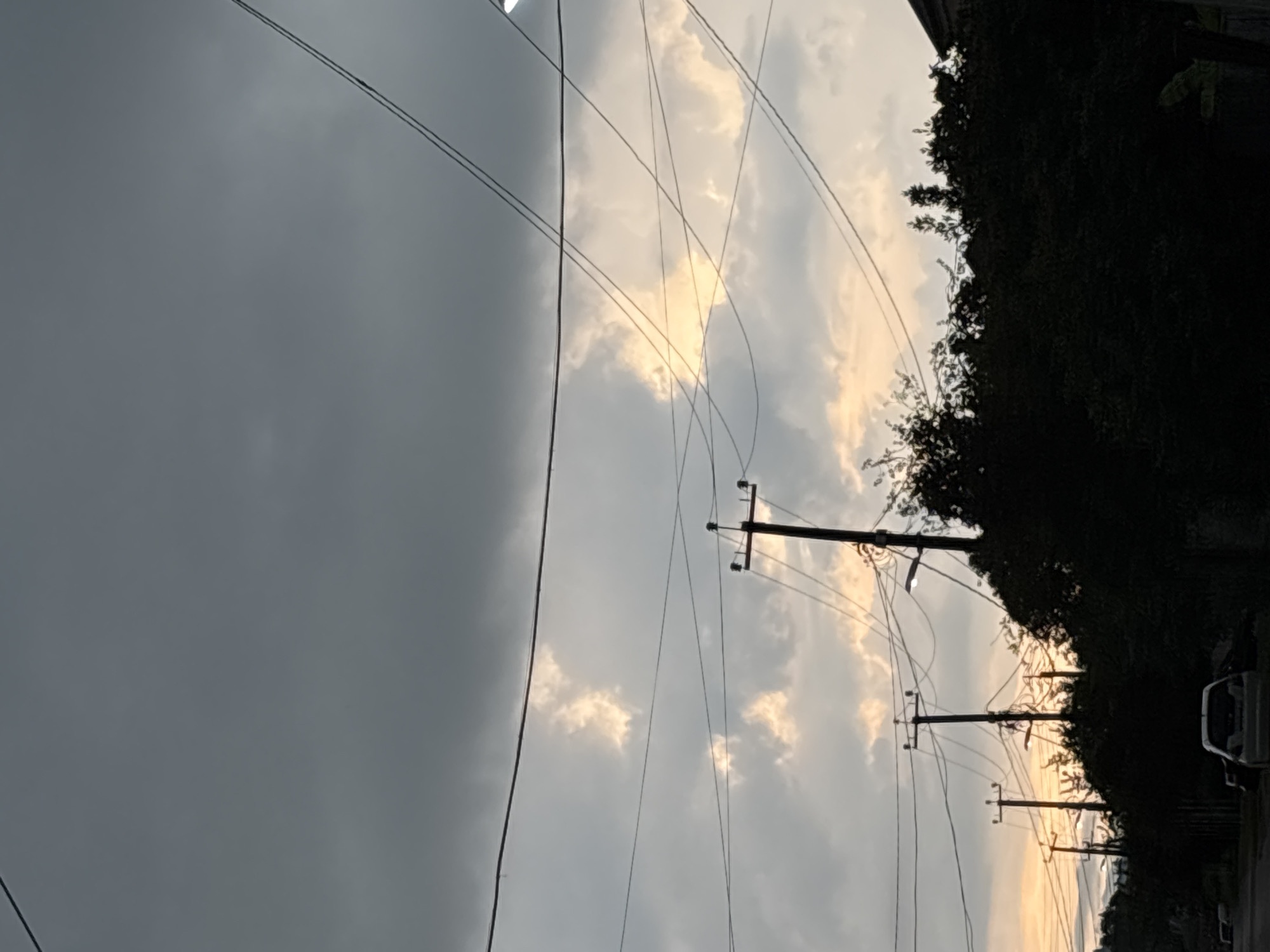 A view of a street lined with trees and utility poles against a stormy sky. The sky is filled with dark, ominous clouds, with patches of yellow light peeking through, suggesting either sunrise or sunset. Multiple power lines crisscross the frame, casting shadows over the scene. Silhouetted against the sky are several utility poles, some with lights attached. The foreground is dominated by dense, dark foliage, with the roofline of a building visible on the right. A white car is parked on the street in the lower left portion of the image. The overall impression is one of a moody, overcast day in a residential or semi-urban area.