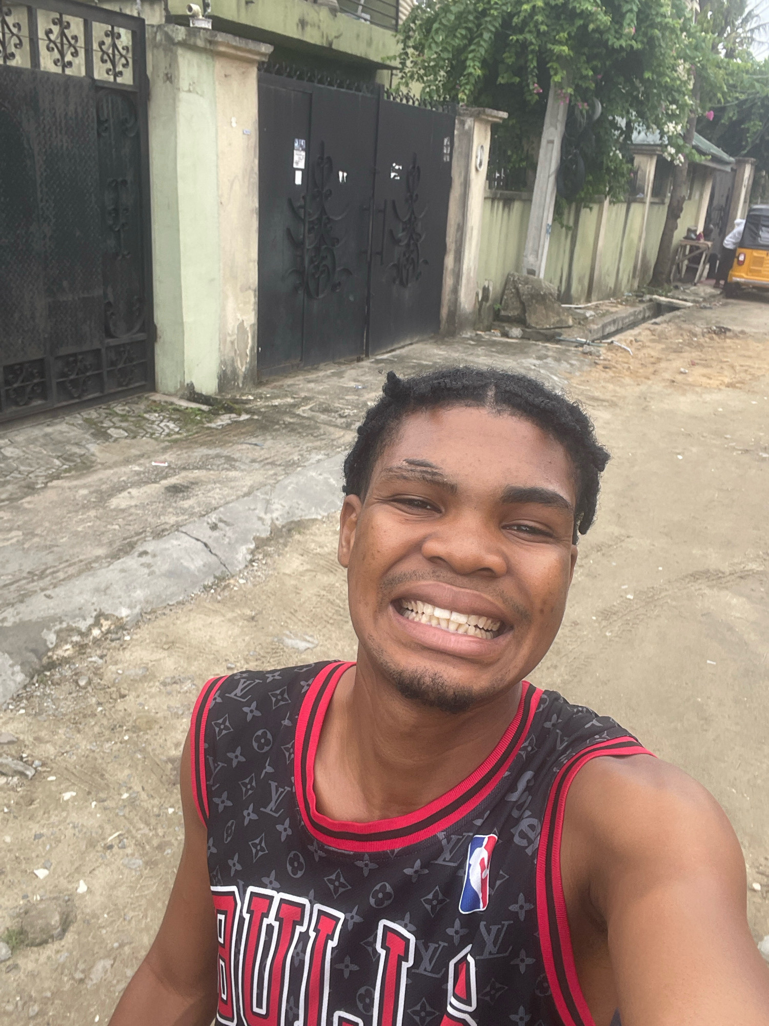 In Ado, Nigeria, a young Black man is captured in a selfie. He is positioned in the foreground, facing the camera with a wide, toothy smile, revealing braces. His braided hair is styled with a middle part. He wears a black sleeveless jersey with a repeating pattern of the Louis Vuitton monogram, accented with red trim and featuring an NBA logo on the left chest. The background depicts a street scene characterized by a dirt and concrete pathway. Behind him, a large, dark metal gate with ornate scrollwork is visible, suggesting a secured property. Adjacent to the gate, a light-colored pillar stands, partially weathered. Further in the background, a fence made of light-colored concrete blocks is lined with lush green foliage. A yellow three-wheeled vehicle is parked on the right side of the image. The overall environment suggests an outdoor urban setting.