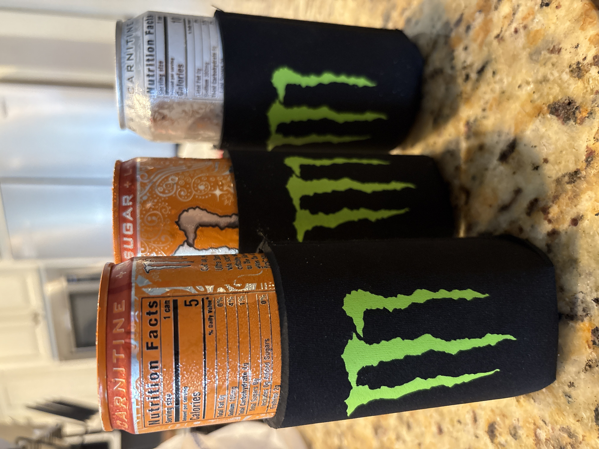 Three Monster Energy drink cans are displayed in black can coolers on a granite countertop. The can coolers have the signature green Monster Energy logo. The can on the left is orange and has "CARNITINE" and "Nutrition Facts" printed on it. The can in the middle is also orange and has "SUGAR + CAFFEINE" printed on it. The can on the right is silver and also has "CARNITINE" and "Nutrition Facts" printed on it. The background shows kitchen cabinets and appliances, suggesting the scene is in a residential kitchen. The lighting indicates it is daytime. The location is Panama City Beach, United States of America.