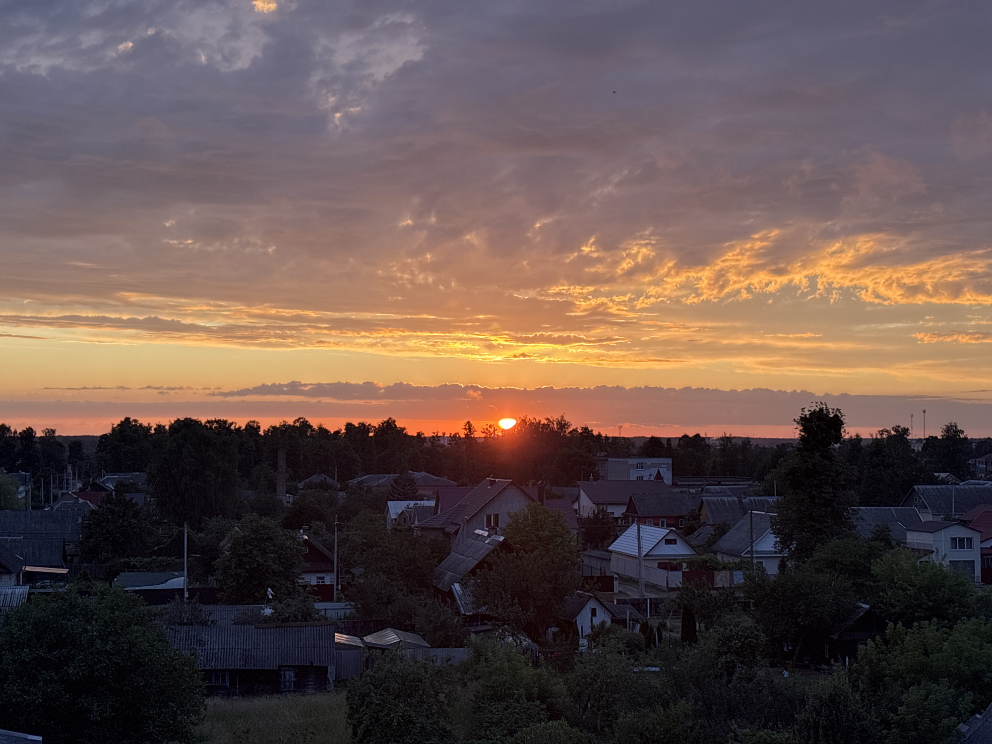 The image depicts a sunset over a village in the Pratasevichy Village Council, Belarus. The sky is filled with colorful clouds, ranging from deep purple to vibrant orange and yellow, as the sun is low on the horizon, casting a warm glow. Below the sky, a dense line of trees forms the edge of a settlement. The village consists of numerous houses with pitched roofs, some appearing older with corrugated metal roofing, while others are more modern. The architecture suggests a rural or suburban setting. The foreground is dominated by dark green foliage, indicating a dense growth of trees and bushes, possibly within gardens or natural areas surrounding the houses. The overall scene is tranquil, characteristic of a rural evening. There are no people visible in the image.