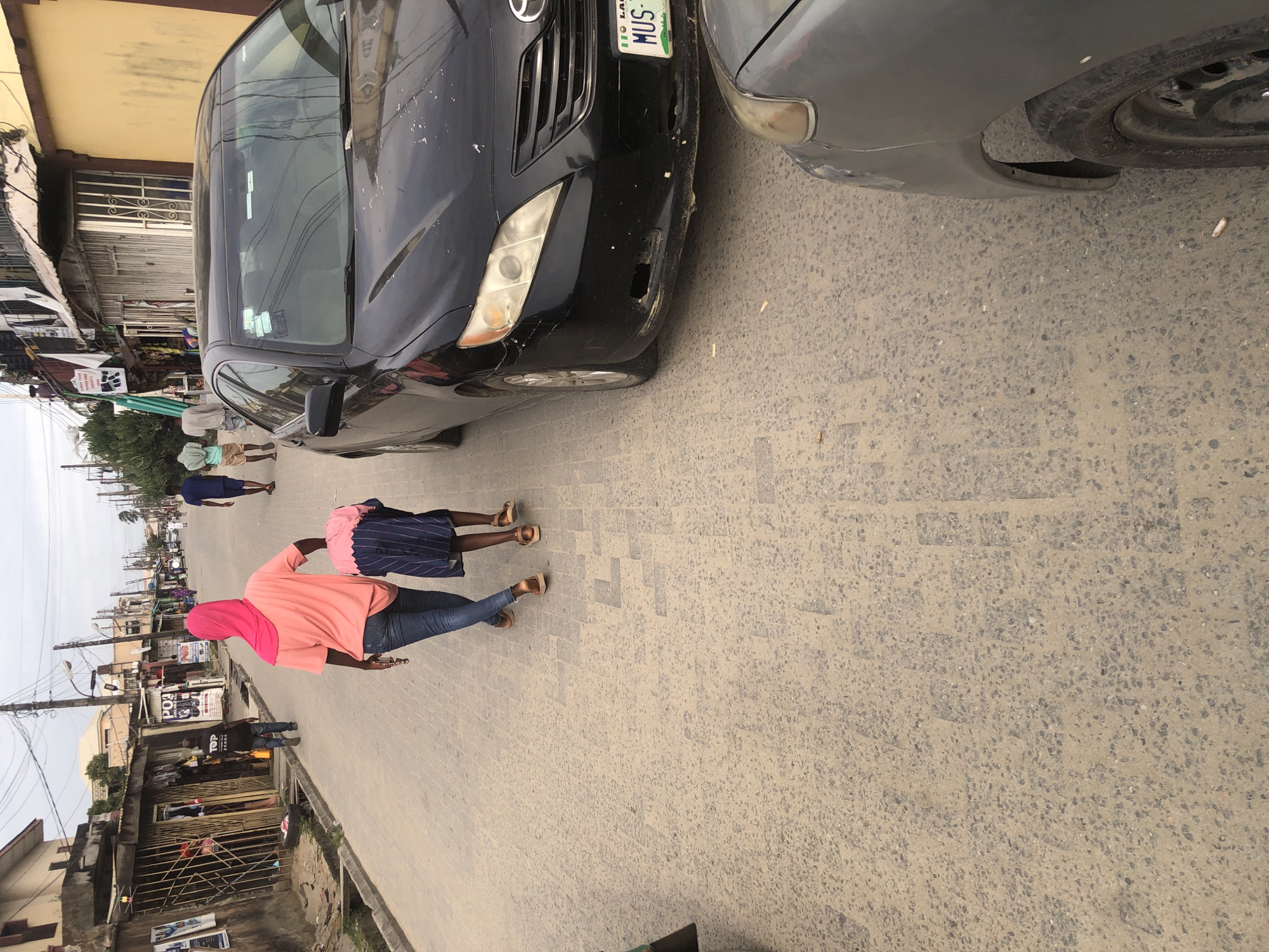 In the city of Ado, Nigeria, a paved street is lined with buildings housing shops and residences. Overhead power lines crisscross the sky. In the foreground, a black Toyota Camry is parked on the right side of the road, with a gray car partially visible in the lower right corner. Two individuals are walking away from the camera on the left side of the street. The person in front is wearing a pink headscarf and a coral-colored t-shirt, with dark jeans and brown sandals. They are carrying a folded blue and white striped garment. Behind them, a smaller individual, presumably a child, is dressed in the garment, which appears to be a blue and white striped dress. Several other pedestrians are visible further down the street, and a man in a black t-shirt with "TOP" written on it is walking away from the camera on the left side. The street is littered with debris.