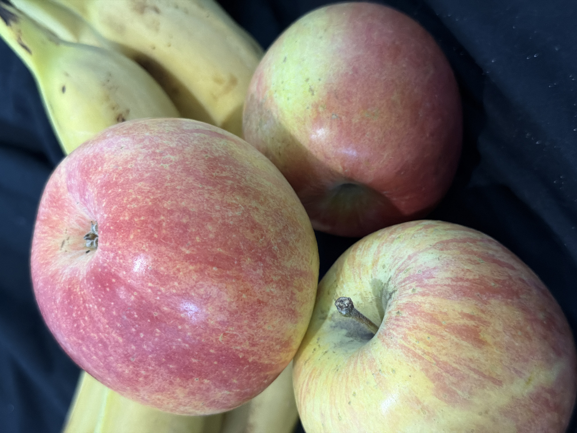 Three apples and a bunch of bananas are displayed against a dark background. The apples are red and yellow with red streaks. The bananas are yellow with some brown spots. The lighting is bright and highlights the textures of the fruit.