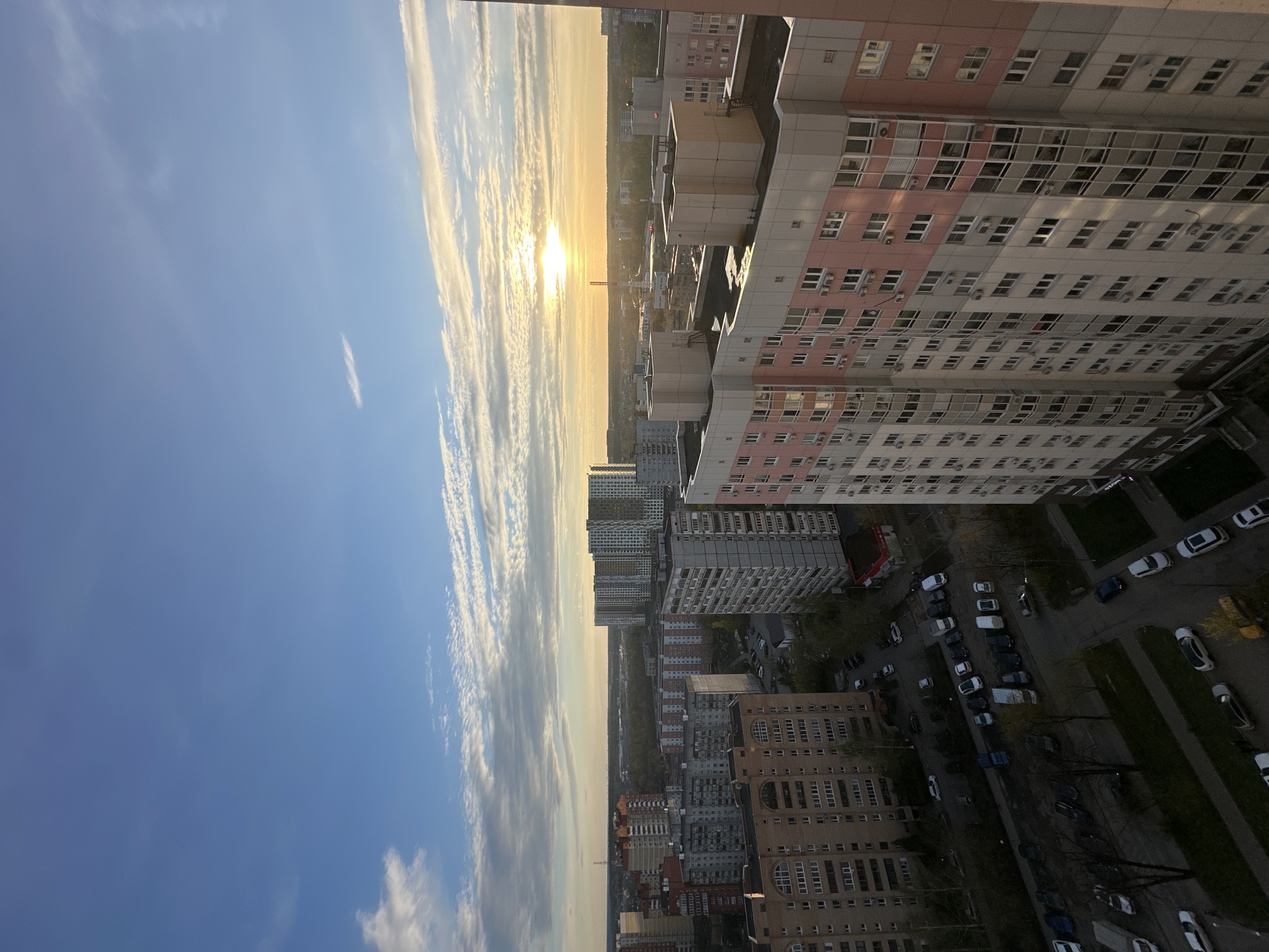 The image depicts a high-angle view over a residential area, likely in Moscow, Russia, given the architectural style. The buildings are predominantly Soviet-era apartment blocks, many with a pink and gray color scheme. The sun is setting, creating a bright line on the horizon and illuminating the sky with a warm glow. Clouds stretch across the sky, adding depth to the scene. Below the buildings, there are streets with parked cars and some greenery. No people are visible in the image. The general environment appears to be an urban residential neighborhood.