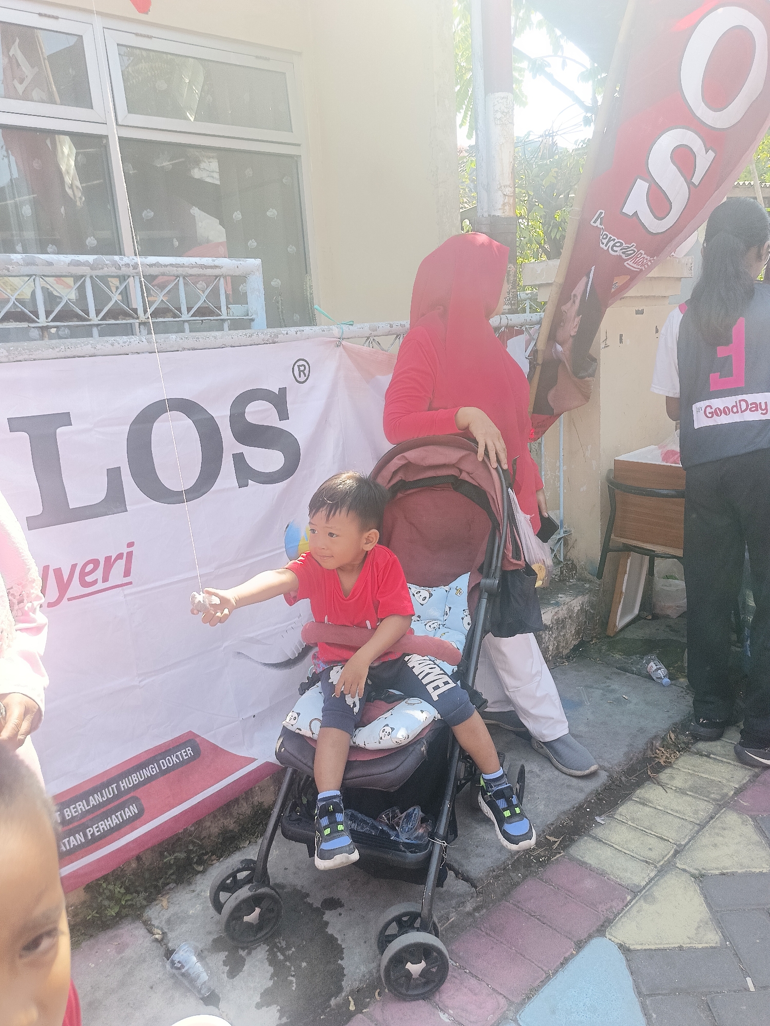 In Gedangan, Indonesia, a young boy in a red t-shirt sits in a stroller. He is holding a string with a small object attached, which he appears to be playing with. A woman wearing a red hijab stands behind him, holding the stroller. To the right, another woman in a black vest with a pink logo is visible, and to the left, partially obscured, is a person in a pink top. A large white banner with black and red text is displayed behind them. The scene takes place outdoors, on a paved area with some greenery visible in the background. A discarded plastic bottle lies on the ground. The lighting suggests it is daytime, with sunlight casting shadows.