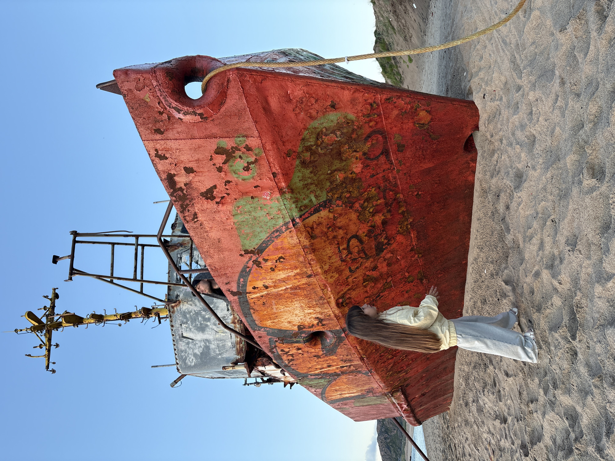 In Teriberka, Russia, a large, rusted red ship rests on a sandy beach. A young girl with long, light-brown hair stands facing the ship, wearing a pale yellow jacket and grey sweatpants. Her right hand is extended towards the vessel. Behind her and to the left, the water is visible, meeting a rocky, treeless coastline. A thick, yellow rope is tied to the ship's bow and trails onto the sand. In the upper section of the ship, a person wearing a black hooded sweatshirt looks upwards. The sky is clear and blue. The ship is covered in graffiti and shows significant wear and tear.