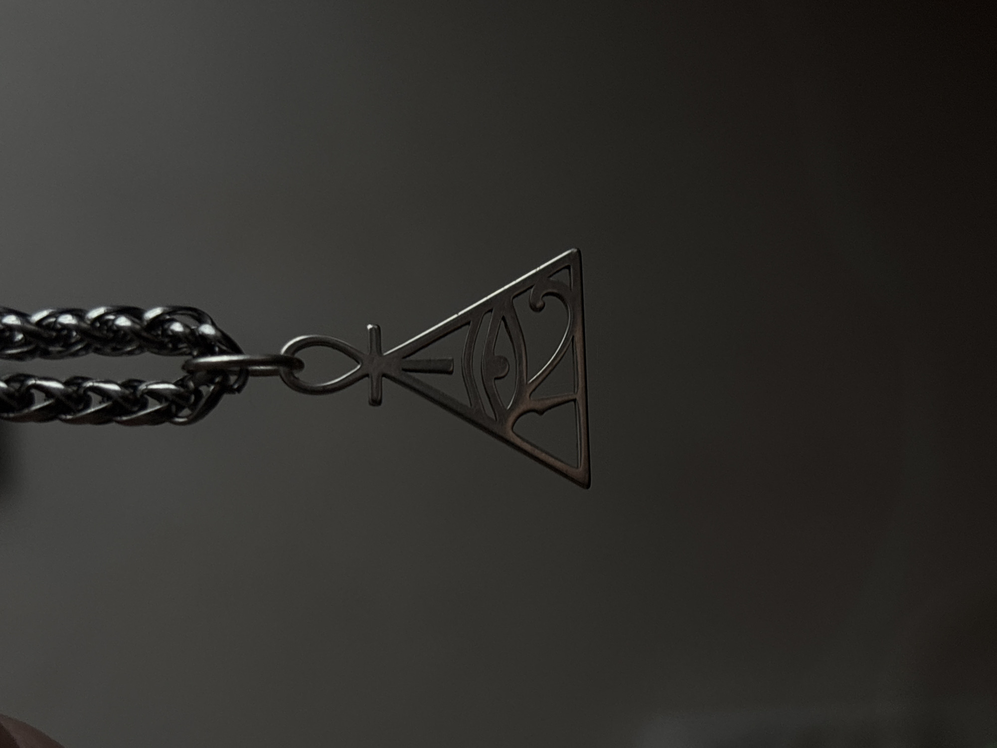 A close-up shot focuses on an ankh symbol on a necklace chain. The ankh, a stylized representation of an eye within a triangular frame, is intricately detailed with swirling patterns inside the eye and a decorative flourish at the top. The necklace chain itself is chunky and made of dark metal, with each link clearly defined. The background is a dark, out-of-focus gradient, suggesting a dimly lit environment. The lighting creates a dramatic effect, with the pendant and chain catching the light, emphasizing their metallic texture and the etched details of the symbol. The scene is still and quiet, with no visible activity other than the presentation of the jewelry. There is no text visible in the image. The overall mood is one of mystery and understated elegance.