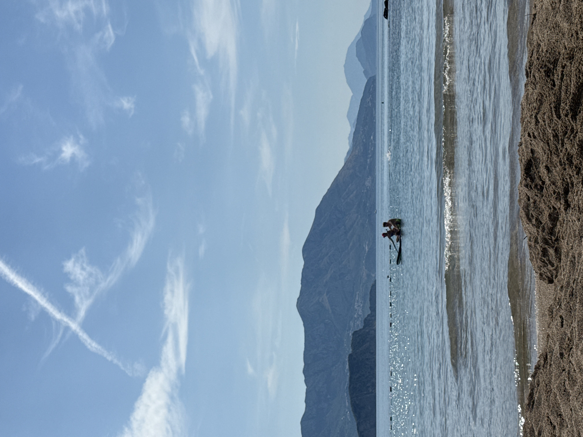 A vertical, outdoor, daytime, medium shot captures a scenic view of the Adriatic Sea and mountainous coastline near Prijevor, Montenegro. In the foreground, the sandy beach transitions into the calm, rippling sea, with small waves lapping the shore. The water is a clear, pale blue, reflecting the bright sunlight, creating a shimmering effect.
Further out in the sea, two individuals are seated on a dark-colored paddleboard. The person in front is wearing a red cap and appears to be holding a paddle. The person behind them is wearing a green swim garment. They are positioned in the middle ground of the image.
Beyond the paddleboarders, the sea extends towards a prominent coastline. On the left, a smaller, darker landmass juts out into the water, with a cluster of buildings visible on its slopes, suggesting a town or village. This is backed by a large, imposing mountain range, its slopes a deep blue-grey, indicating distance. The mountains form a dramatic backdrop, their peaks and ridges clearly defined against the pale blue sky.
The sky itself is vast and bright, with scattered, wispy white clouds. A distinct, arrow-shaped contrail, likely from an airplane, stretches across the upper portion of the sky, adding a touch of man-made presence to the natural landscape. The overall impression is one of a peaceful, sunny day at the beach, with a blend of natural beauty and distant signs of human habitation.