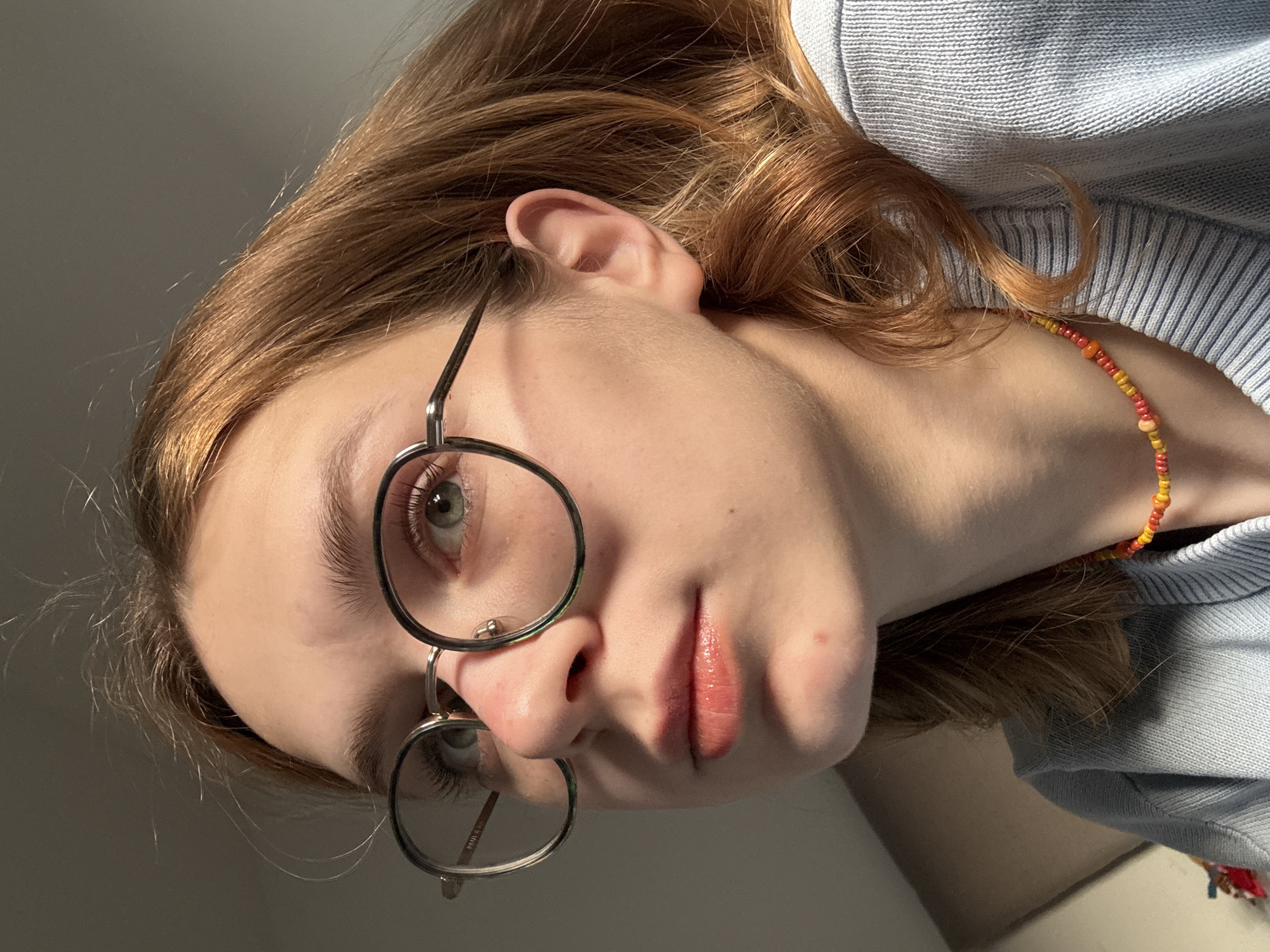The primary image is a close-up portrait, rotated 90 degrees clockwise, depicting a young individual indoors. The subject has reddish-brown, wavy hair and striking light green-blue eyes that look slightly upwards. They are wearing round, thin-framed metal glasses, with the brand name "PRADA" subtly visible on the inside arm. Their lips are a light pink, slightly parted, with a hint of gloss.
The person is dressed in a light blue, ribbed knit sweater. A vibrant, multi-colored beaded bracelet, featuring hues of red, orange, and yellow, adorns their wrist. Strong, directional sunlight illuminates the subject's face and hair from the left, creating distinct highlights and shadows, suggesting a bright window or light source, likely indicating morning or late afternoon. The background is an indistinct, plain light grey surface. The expression is neutral and calm. The setting is an indoor space, within the location context of Kragujevac, Serbia.