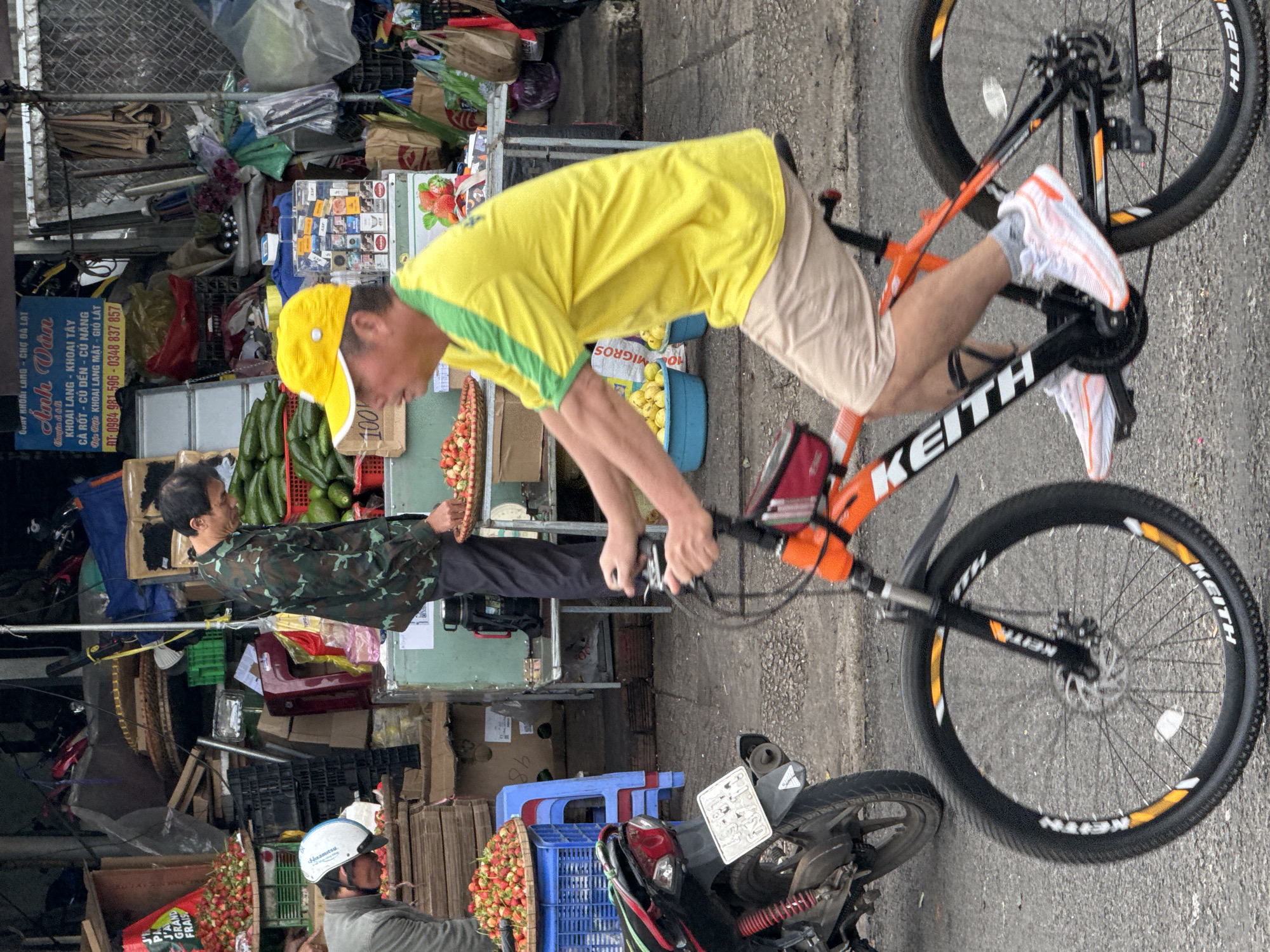 In Da Lat, Vietnam, a man wearing a yellow cap and a bright yellow shirt with green trim is cycling on a street. He is also wearing light-colored shorts and white athletic shoes with orange accents. The man's bicycle is orange and black, with the brand name "KEITH" visible on the frame and wheels. In the background, a market stall is set up with various fruits, including strawberries and avocados, displayed in baskets and crates. A motorcycle is parked to the left of the cyclist, and other market stalls with goods and signs are visible further back. Another person, wearing a camouflage jacket, is seen standing at one of the market stalls. The overall environment appears to be a busy street market.