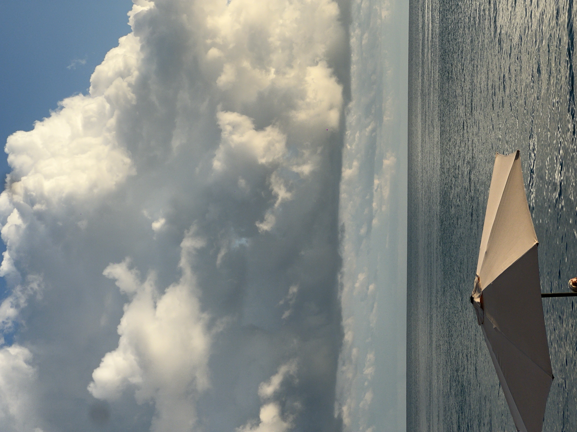 A wide, off-white umbrella is positioned in the foreground, angled towards the right side of the frame, casting a shadow. Behind the umbrella, a calm, dark blue body of water stretches to the horizon. The sky above is filled with dramatic, dense, grey and white clouds, with patches of blue sky visible. The image appears to be taken at a beach or seaside location, potentially in Sokhumi, Abkhazia, given the presence of the sea and the characteristic cloud formations often seen in coastal areas. The overall mood is somewhat overcast and atmospheric.