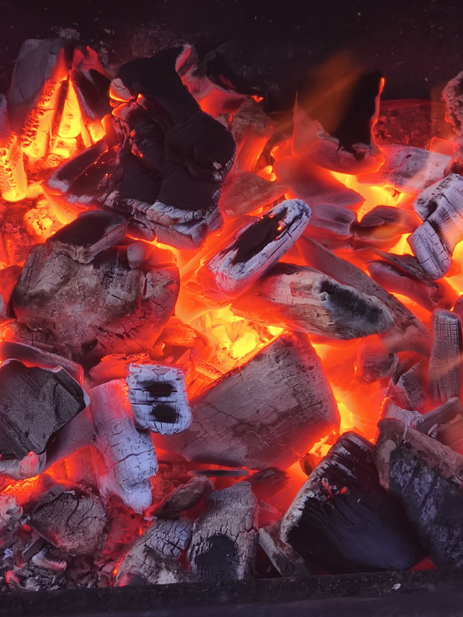 The image shows burning wood in a grill. The wood is glowing red and orange, with some pieces still retaining their original gray color. The composition fills the frame, suggesting a close-up perspective. The location is not discernible from the image.