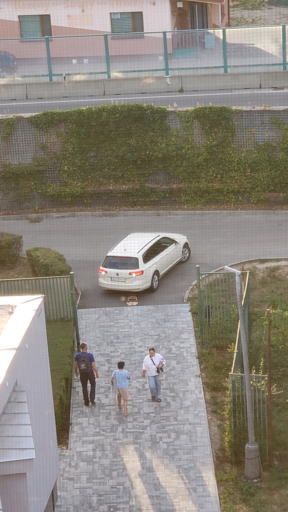 The image is an outdoor, high-angle shot of a paved walkway adjacent to a road. On the road, a white station wagon is parked. Three people are walking on the paved walkway, moving from the bottom of the frame towards the top. The person furthest to the left is wearing a blue t-shirt and dark pants, carrying a backpack. The middle person is wearing a light blue t-shirt and light-colored shorts. The person furthest to the right is wearing a white shirt and blue jeans, carrying a bag over their shoulder. The environment includes some green foliage and a fence. The scene is consistent with a residential area in the city of Prešov, Slovakia.