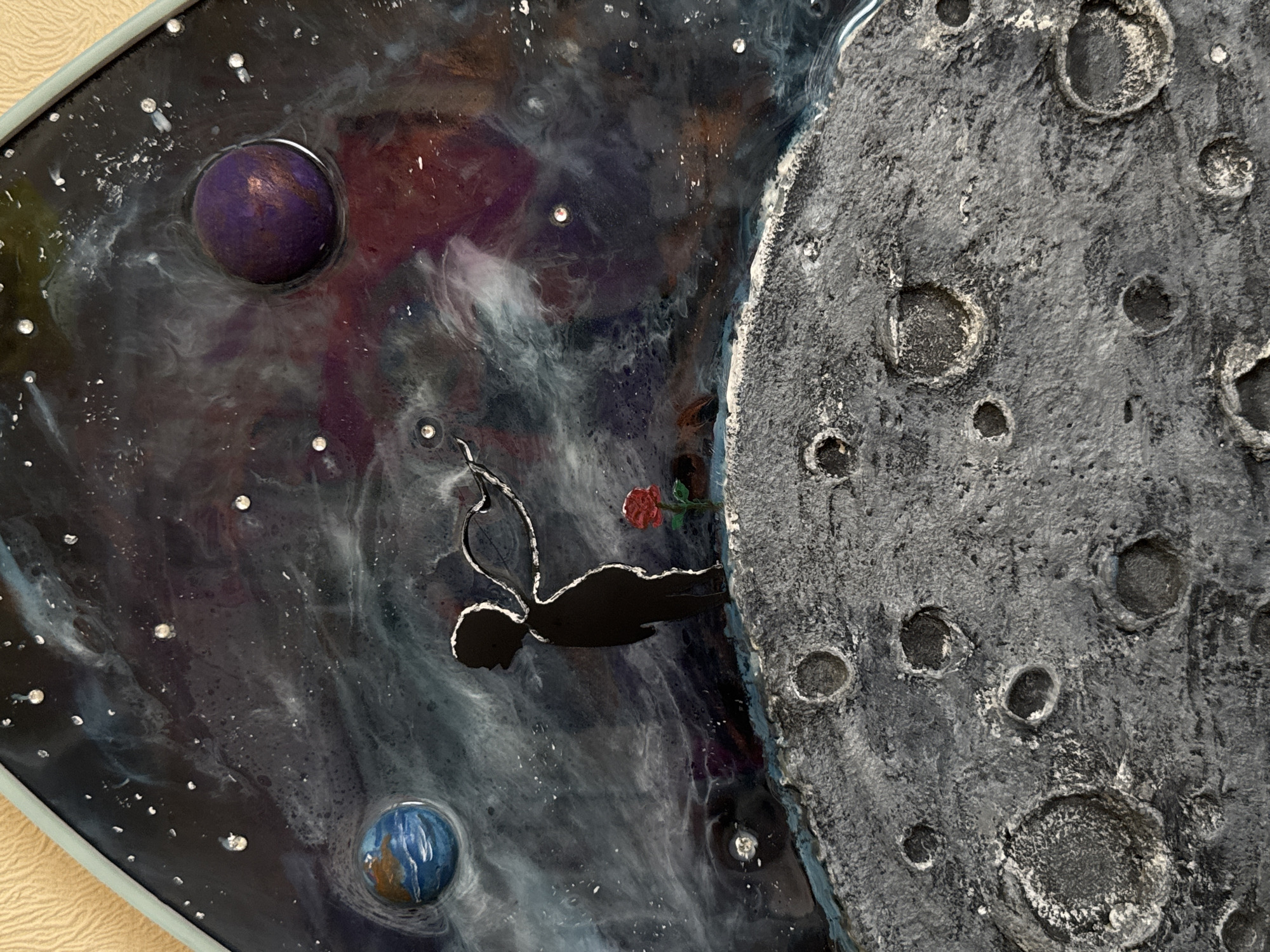 The image depicts a resin art piece inspired by "The Little Prince". A silhouette of a child with wings stands on a textured, cratered surface resembling the moon. To the child's right is a single red rose. In the background, a swirling nebula of dark blues, purples, and white occupies the space, with small, sparkling "stars" scattered throughout. Two planets are visible: one blue and green, resembling Earth, and another spherical, purple planet. The art piece is framed by a light blue border. There is no indication of the city of Kolomna, Russia.