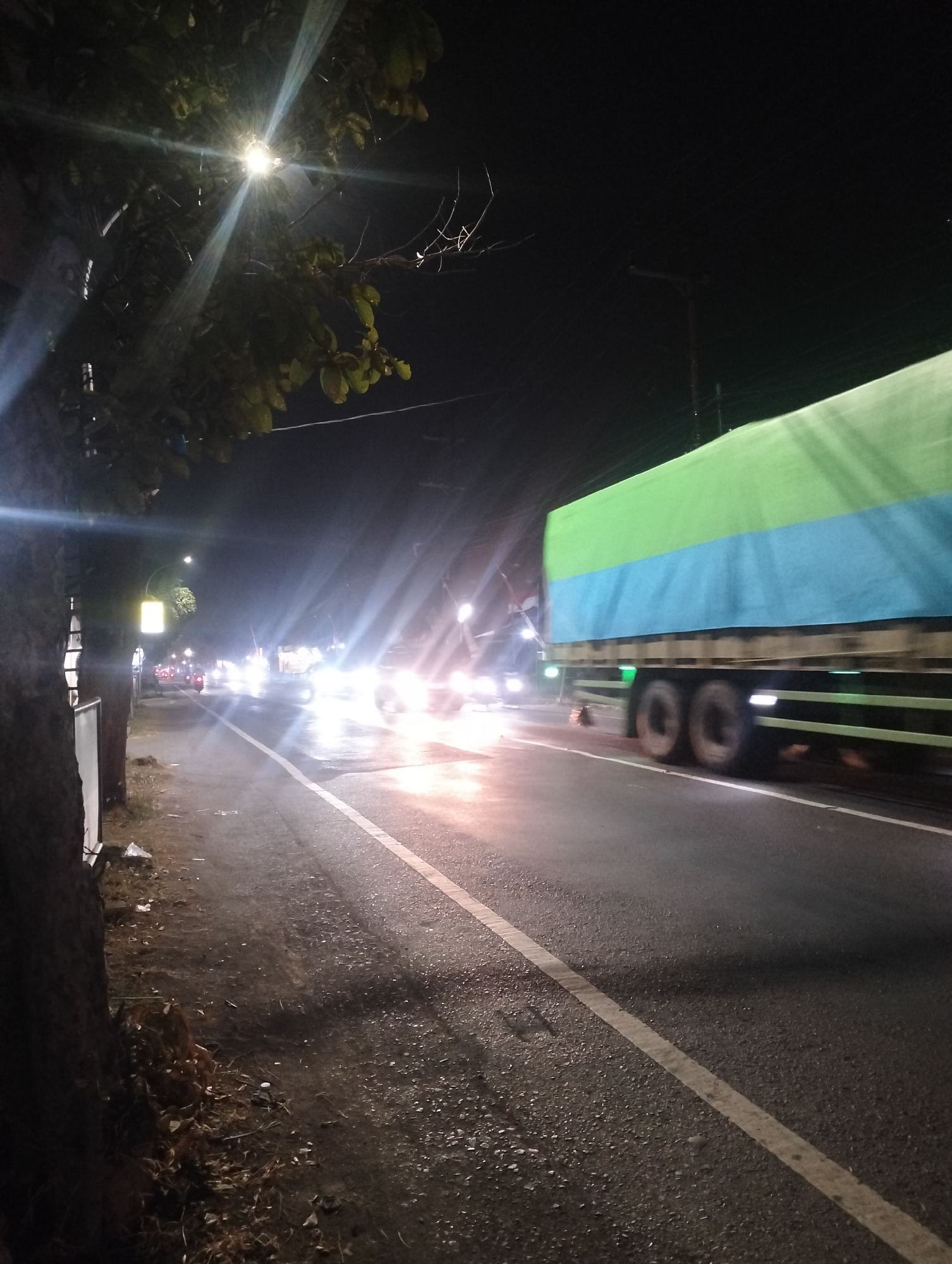 The image depicts a nighttime scene on a street in Driyorejo, Indonesia. A large semi-trailer truck with a green and blue tarp-covered cargo is driving on the right side of the road, moving from left to right. The truck is illuminated by streetlights and its own headlights. To the left of the road, a sidewalk area is visible with a tree and some scattered debris. Further down the road, other vehicles with their headlights on are approaching, creating significant light glare. A streetlamp with a yellow fixture is also visible on the left side of the road, illuminating a section of the sidewalk and the tree. The road surface appears to be asphalt and is wet, reflecting the lights from the vehicles. The sky is dark, indicating it is nighttime.