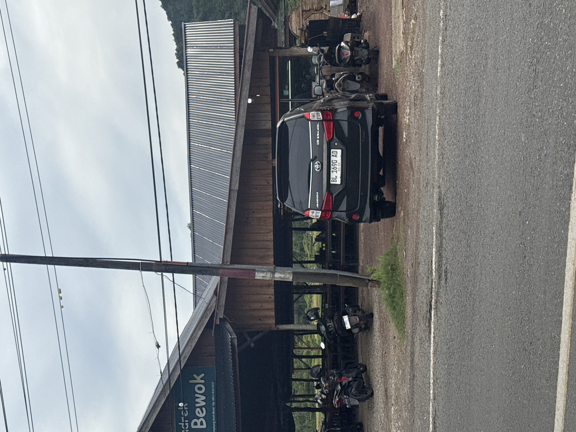 The image depicts a roadside scene in Lampisang, Indonesia, likely in front of a restaurant or food stall. A black Toyota Avanza with license plate BL 1690 AD is parked in the foreground. Behind the car, a rustic, open-sided building with a corrugated metal roof is visible. Several motorcycles are parked in front of the building, suggesting customers. The overall environment appears to be rural or semi-rural, with a dirt or gravel lot surrounding the building and a paved road in the foreground. A utility pole stands near the building. In the background, a tree-covered hill is visible. The sky is overcast.