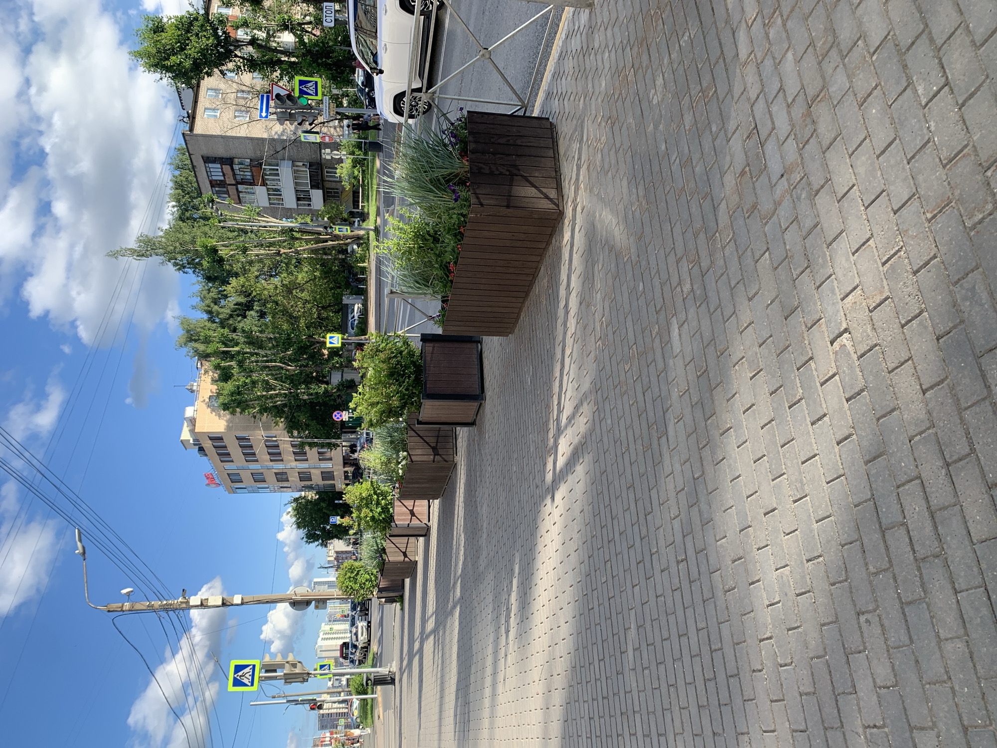 This is a photograph taken on a sunny day, possibly in the afternoon, showing a street in the city of Perm, Russia. The foreground is dominated by a gray brick sidewalk, with a clear pattern of rectangular bricks. In the mid-ground, a series of large, dark brown wooden planter boxes are arranged along the edge of the sidewalk. These planters contain various green plants, including tall grasses and smaller leafy shrubs, with some small red and purple flowers. A metal railing separates the planters from the street.
The street itself is visible, with traffic. A white SUV is parked on the right side of the street. To the left of the planters, a pedestrian crossing is marked on the road. Traffic lights and street signs are visible. One sign is a yellow diamond shape with a black pedestrian symbol, indicating a pedestrian crossing. Another sign is a blue square with a white "P", indicating parking. Further down the street, several cars are seen moving.
In the background, there are multi-story buildings. One building on the left appears to be a light-colored commercial building with red signage. Another building on the right is a residential building constructed of brick. Trees line the street between the buildings and the sidewalk. The sky is blue with scattered white clouds. The overall impression is of an urban street scene with landscaping elements designed to beautify the area.