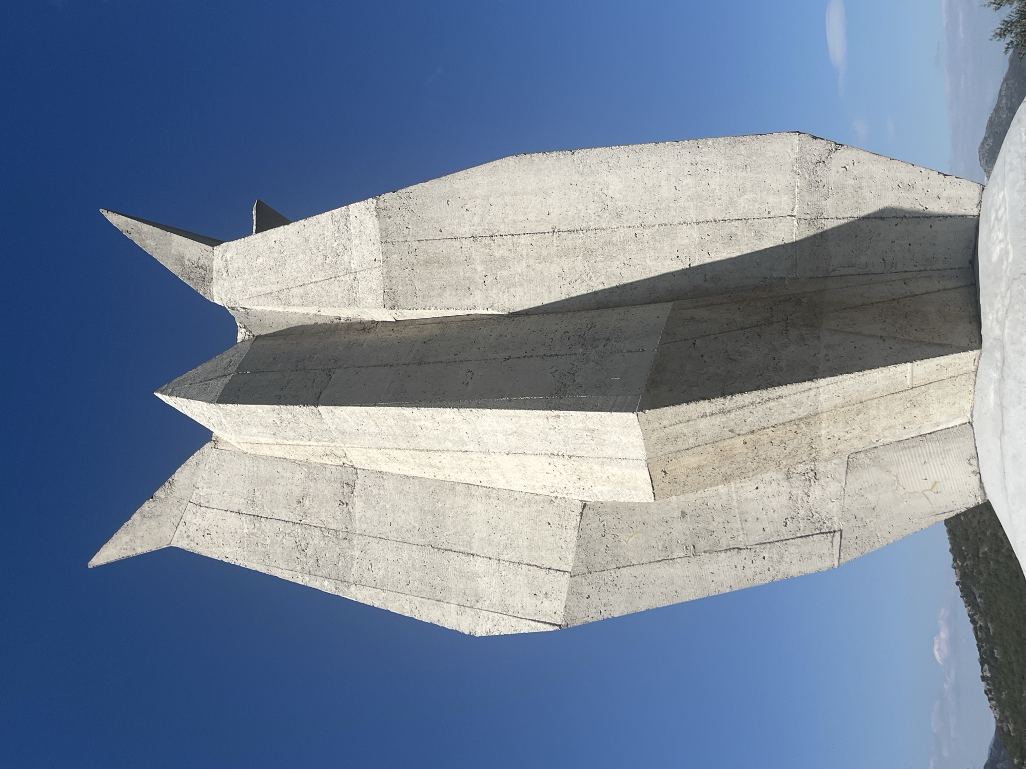 The image shows a concrete monument or sculpture in Godinje, Montenegro. The sculpture is composed of stacked, angular concrete segments, forming a sharp, geometric shape against a clear blue sky. The concrete surface shows signs of weathering and exposed aggregate. Part of the monument abuts a whitewashed element that includes vegetation from surrounding hills on the right side of the frame.