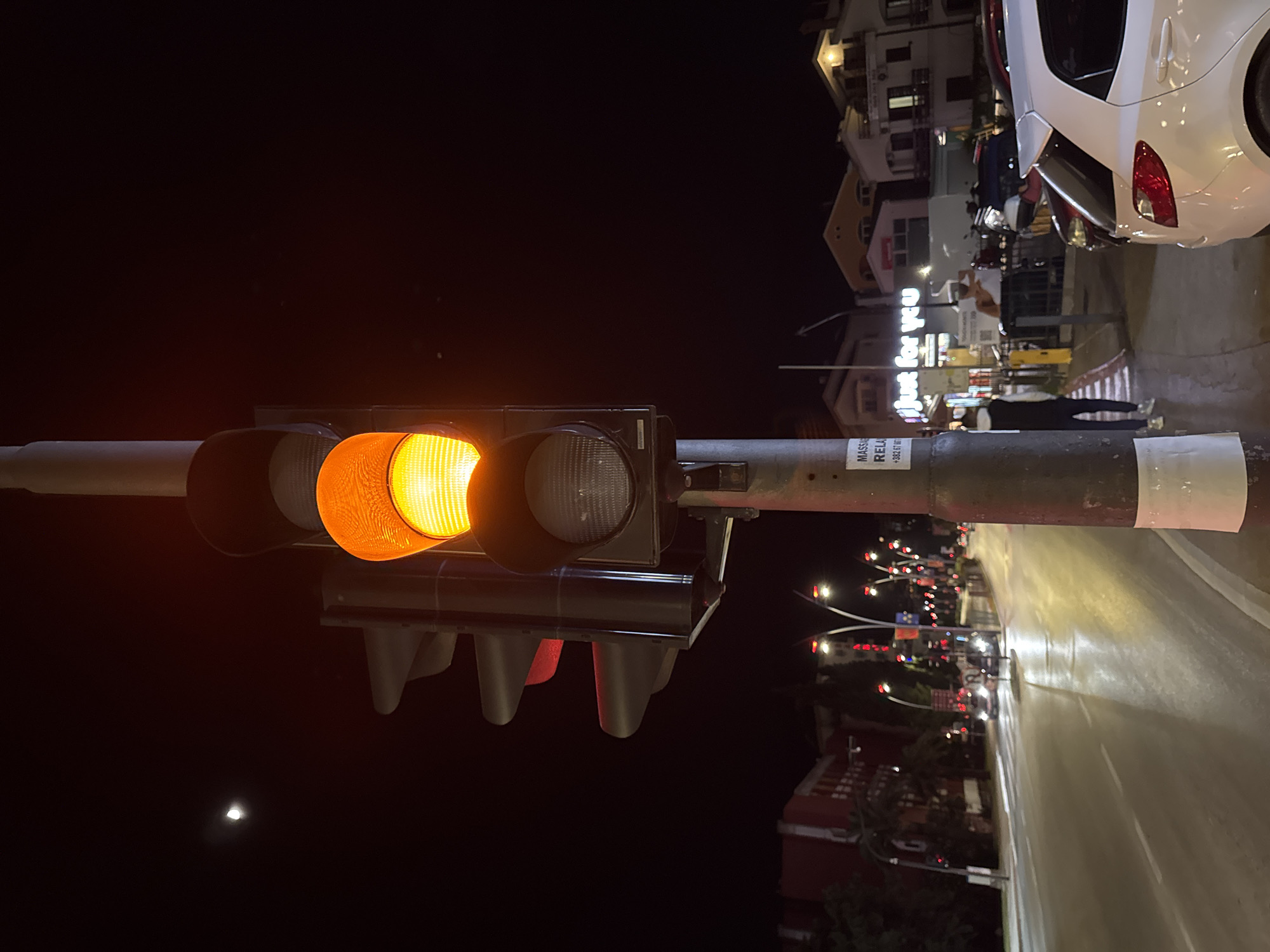 The image depicts a nighttime scene at an intersection in Budva, Montenegro. A traffic light is prominent in the foreground, with the amber light illuminated. The pole supporting the traffic light has a sign reading "MASSA RELA". In the background, city lights illuminate buildings and a street. A white car is parked on the right side of the image. Pedestrians are walking near shops with the words "Just for you" visible. The road is wet, reflecting the city lights. The sky is dark, with a sliver of the moon visible. Other traffic lights are seen further down the street.