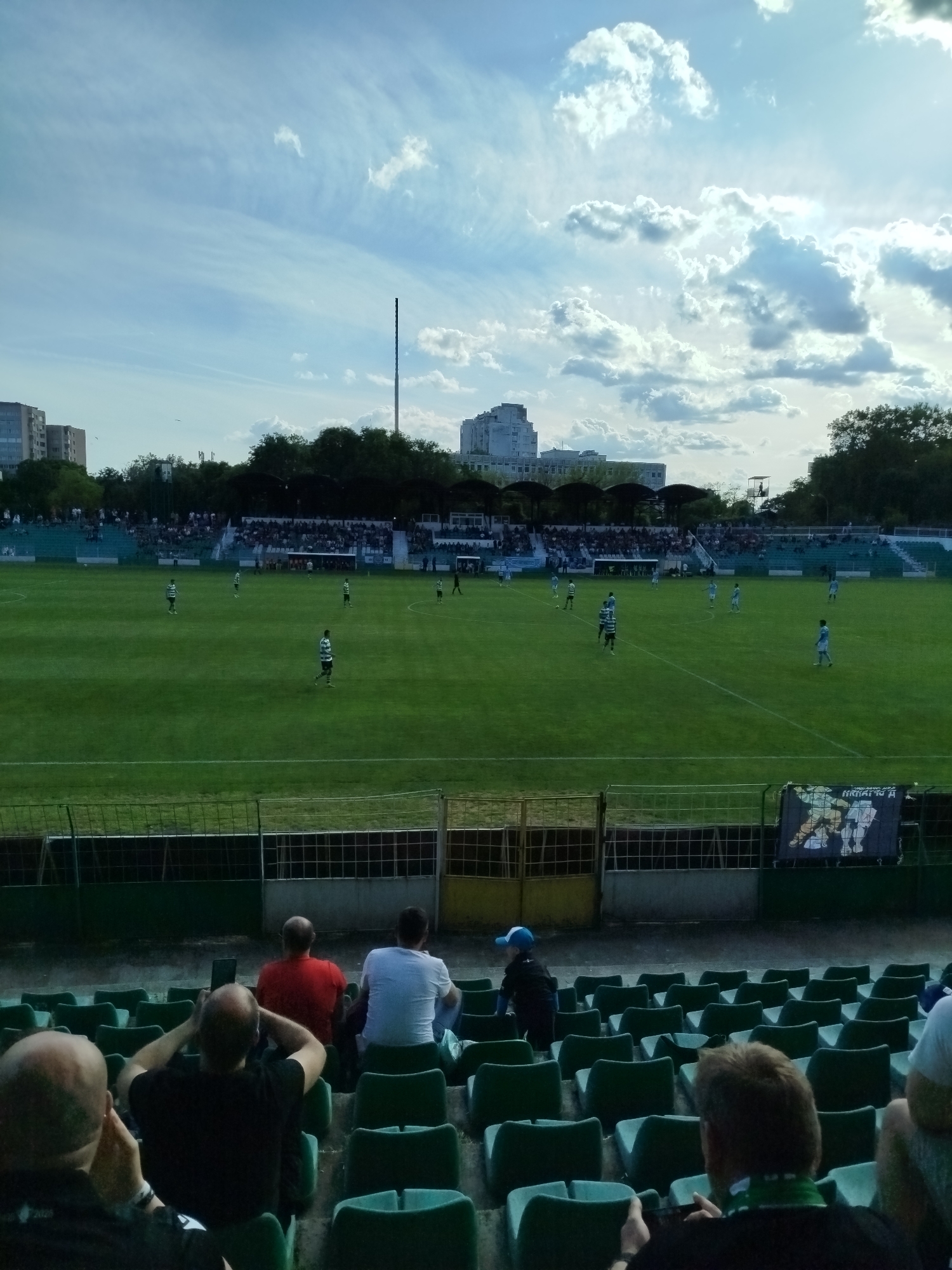 The image depicts a soccer game in progress at a stadium in Varna, Bulgaria. The green field is visible, with players in green and white uniforms distributed across the field. Spectators are seated in the stands, and other spectators are sitting in green seats closer to the camera. The sky is partly cloudy. A tall metal pole stands behind the field. A building is visible in the background, situated above the tree line behind the stands. A banner with the inscription "KAPATOH A" hangs above the seats. In the foreground, several people sit in green stadium seats, watching the game. The seating area appears to be partially filled.