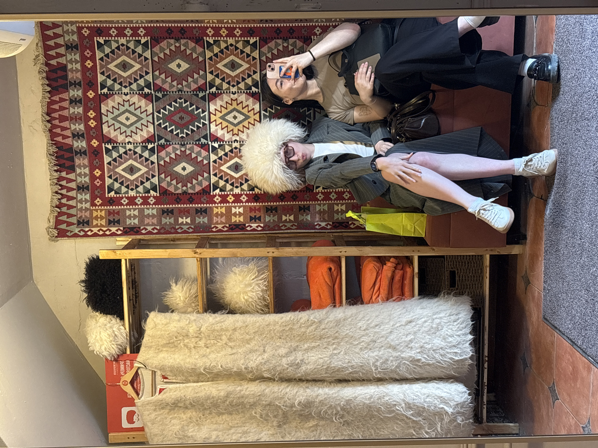 Two people are lying on a rug. One is taking a photo of the other who is wearing a large white hat and striped suit. A shelf unit is beneath the rug, filled with white and orange items. The location is likely a shop in Perm, Russia, possibly a clothing or textile store.