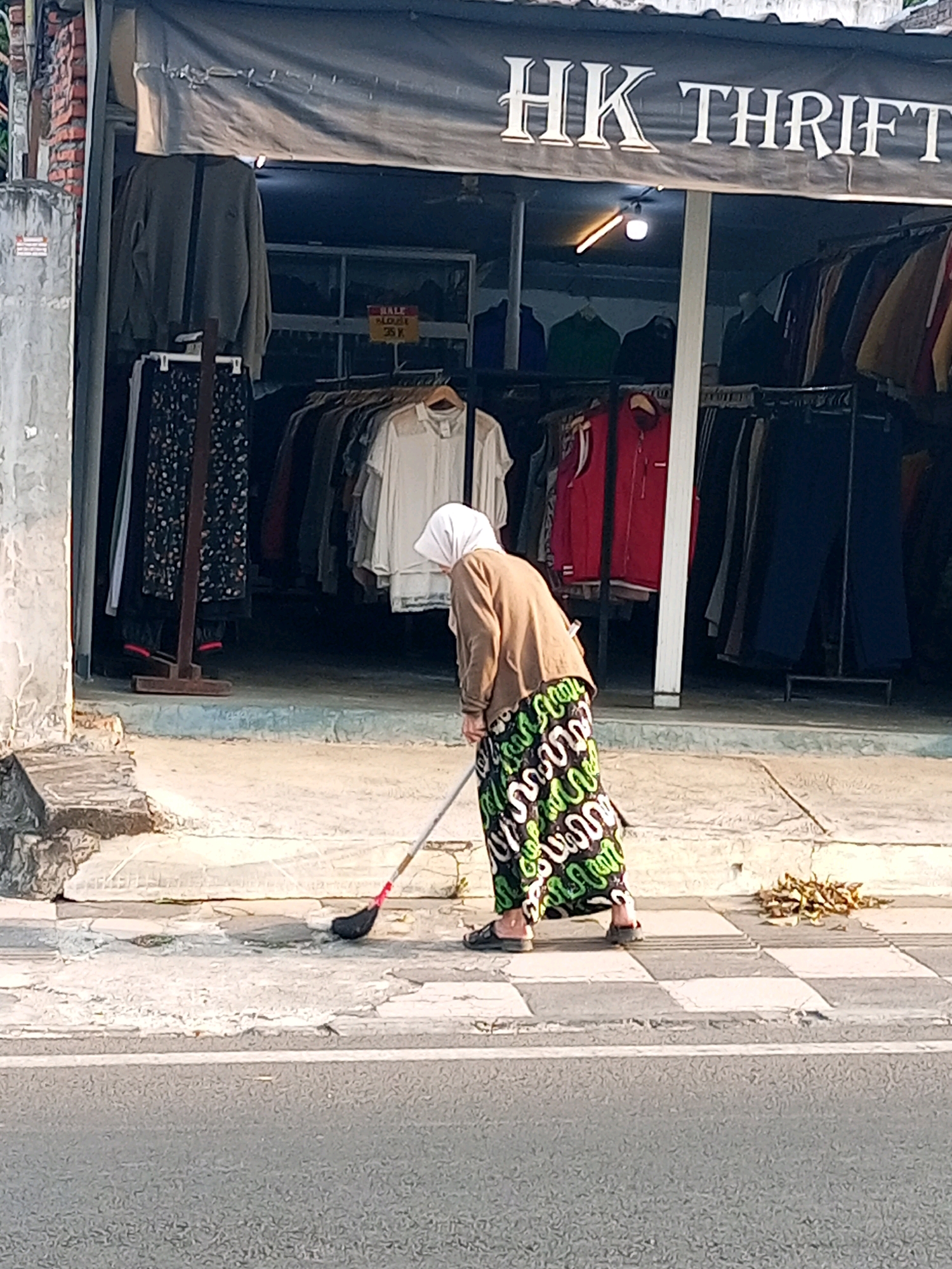 An elderly person wearing a white hijab, a brown cardigan, and a colorful patterned skirt sweeps the sidewalk in front of a thrift store with the sign "HK THRIFT" in Batu, Indonesia. The store displays various clothing items on racks, including shirts, jackets, and pants. The sidewalk is made of concrete and has a crosswalk pattern. A pile of dry leaves is visible on the right side of the sidewalk. The scene is captured during daytime, with sunlight casting shadows.