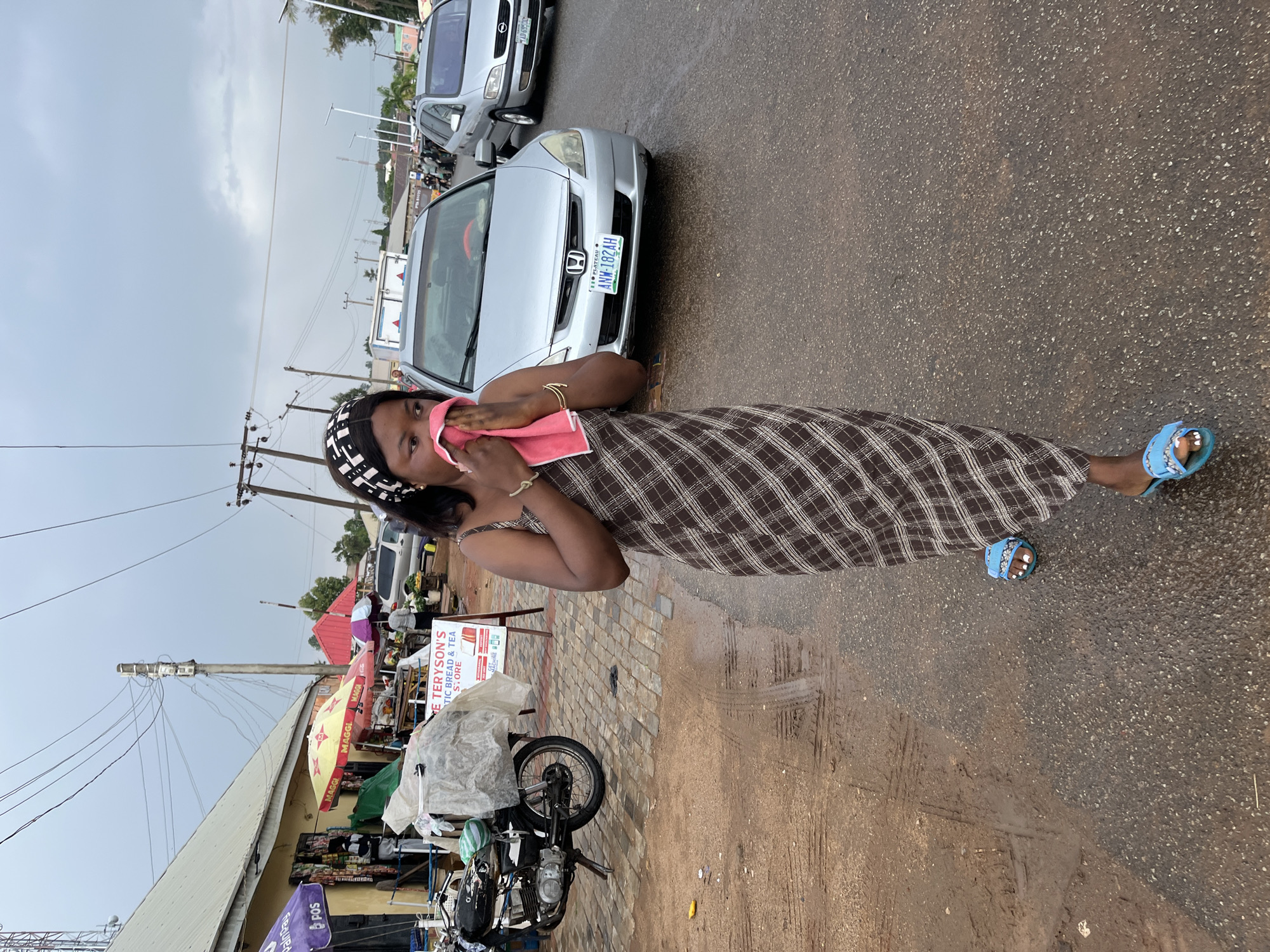 A woman stands on a street in Jos, Nigeria, holding a pink cloth to her face. She is wearing a brown plaid dress and blue sandals. In the background, there are cars, motorcycles, and shops with umbrellas. A sign reads "E TERYSON'S ARTIC BREAD & TEA STORE." A silver car has the license plate "ANW-182AH." The street appears wet.
