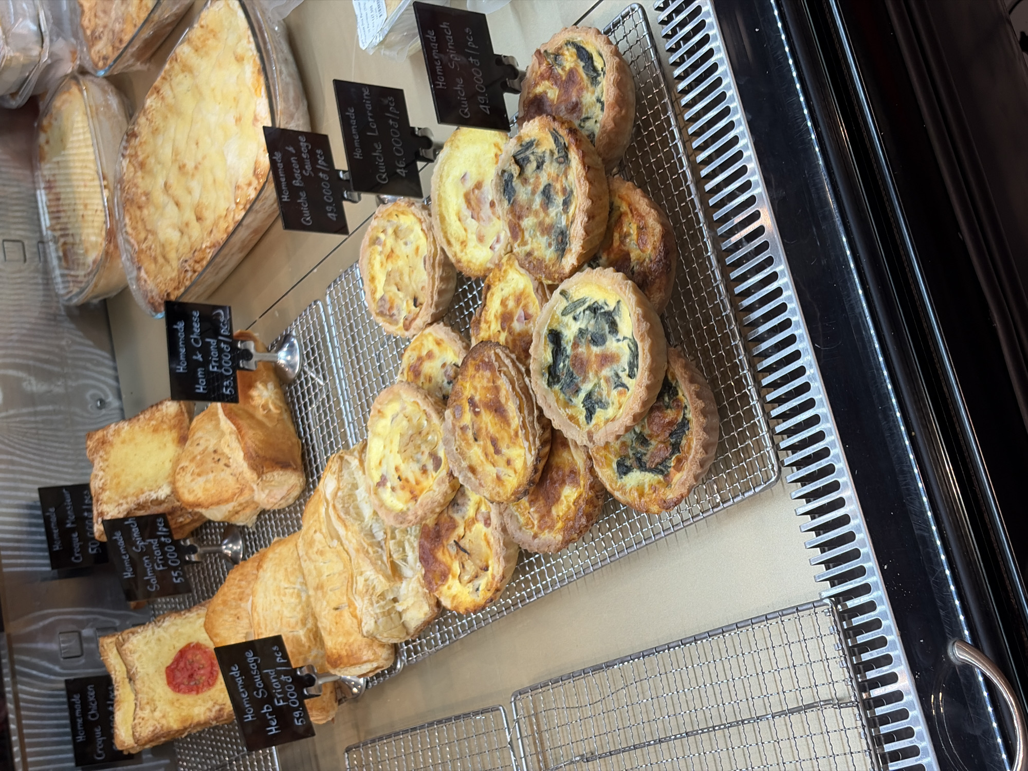 This image depicts a refrigerated display case within a commercial establishment, likely a bakery or deli, located in Thủ Đức, Vietnam. The case showcases various baked goods and prepared meals arranged on metal wire racks.
In the upper left, two "Homemade Croque Chicken" items are visible, one featuring a round red tomato slice on its golden pastry crust. Adjacent to these are a "Homemade Croque Monsieur" priced at 50,000₫/pcs and a "Homemade Salmon Spinach Friand" priced at 55,000₫/pcs, both rectangular or square pastries. Below them, a row of elongated puff pastries identified as "Homemade Herb Sausage Friand" are priced at 59,000₫/pcs. A partially visible "Homemade Ham & Cheese Friand" sign indicates a price of 53,000₫/pcs.
In the middle-right section of the display, two large rectangular glass dishes contain baked items with a browned, cheesy topping, resembling lasagna or a baked pasta dish. One is in the foreground, and another is positioned behind it.
The foreground displays a collection of multiple small, round quiches arranged on a wire rack. These include "Homemade Quiche Bacon & Sausage" at 49,000₫/pcs, "Homemade Quiche Lorraine" at 46,000₫/pcs, and "Homemade Quiche Spinach" at 49,000₫/pcs. The quiches exhibit varied toppings, including spinach, bacon, sausage, and onions.
All food items are presented with small, black rectangular signs on metal stands, displaying their names as "Homemade" products and their prices in Vietnamese Đồng (₫) per piece. The display case features a reflective dark surface at the bottom with a visible LED light strip. No individuals are present in the frame.