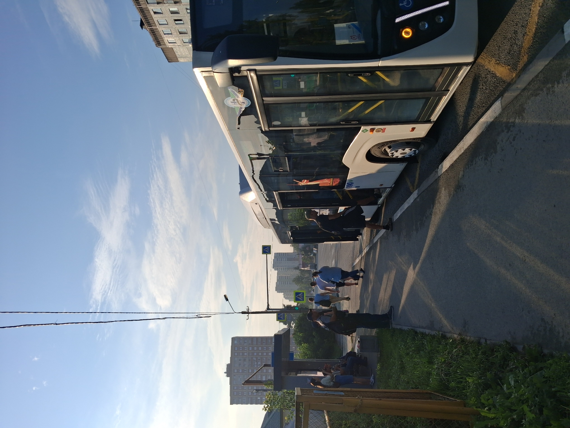 This image captures a street scene in Novosibirsk, Russia, near a bus stop. A white bus with black-tinted windows is stopped at the curb, with its doors open. Several people are visible, some waiting at the bus stop and others boarding or disembarking the bus.
At the bus stop, a woman in a blue skirt and white top is standing, while two other individuals are seated on a bench. A man wearing a dark shirt and backpack stands nearby, looking towards the bus. Further down the sidewalk, a group of people, including a man in a light blue shirt and a woman in a denim jacket, are walking away from the bus stop. Another man in a dark shirt and shorts is partially visible entering the bus.
The environment suggests a late afternoon or early morning, with long shadows cast across the asphalt. The sky is partly cloudy with a light blue hue. In the background, several multi-story apartment buildings are visible, characteristic of urban settings. A bus stop shelter with a chain-link fence is also present on the left side of the image. Traffic signs, including pedestrian crossing signals, are visible on poles along the road. The overall scene depicts a typical urban transportation hub.