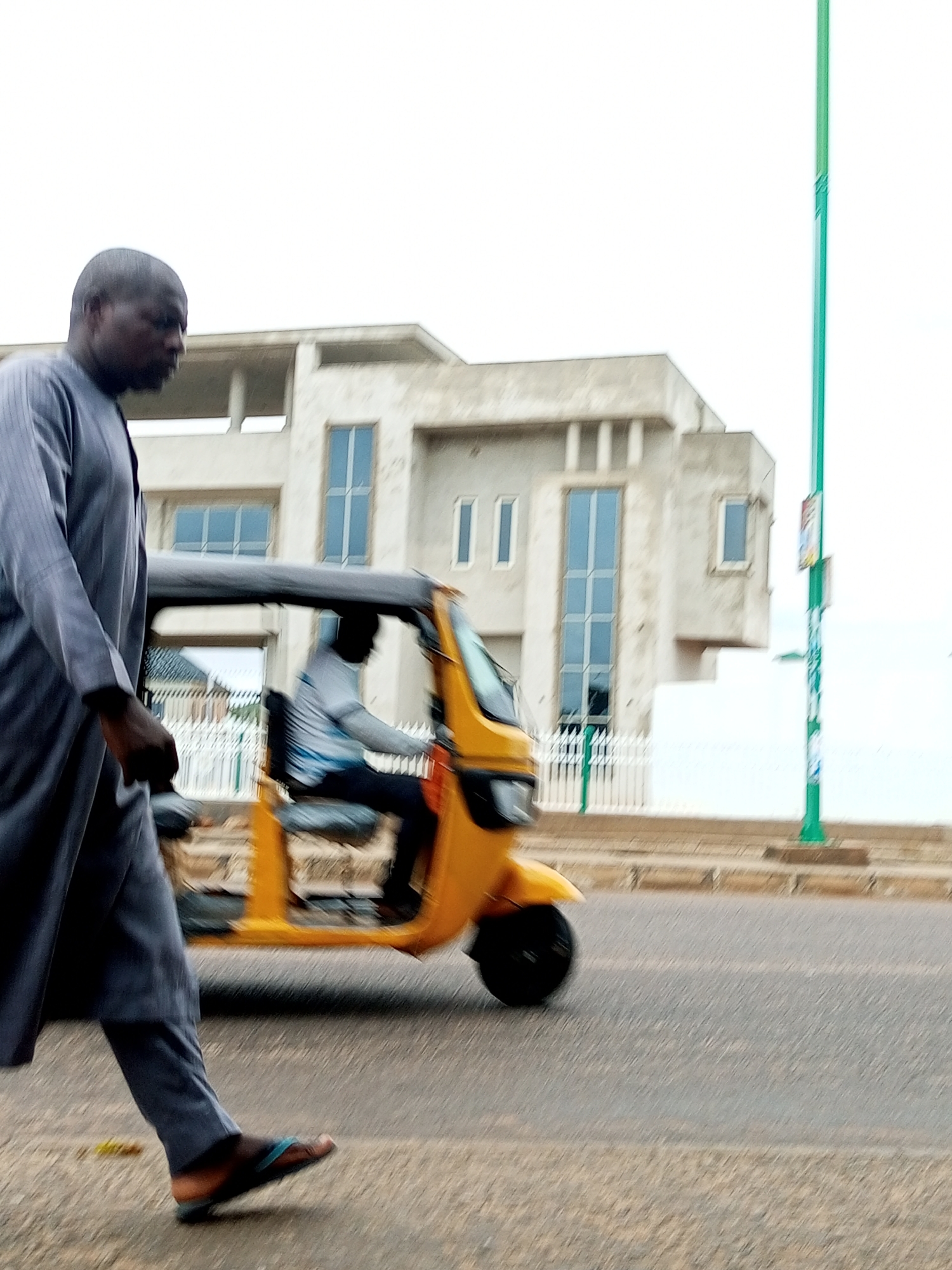 The image depicts a street scene in Pantame, Nigeria, featuring a man walking in the foreground and a yellow auto-rickshaw moving in the background. The man on the left is wearing a grey traditional garment and flip-flops, walking on a paved road. To his right, a yellow auto-rickshaw is in motion, with a driver in a white shirt visible inside. Behind the auto-rickshaw, a modern building with large windows and a white fence is partially visible. A green pole, possibly a lamppost or street sign, stands tall on the right side of the image. The sky is overcast. The overall impression is one of everyday life on a Nigerian street.