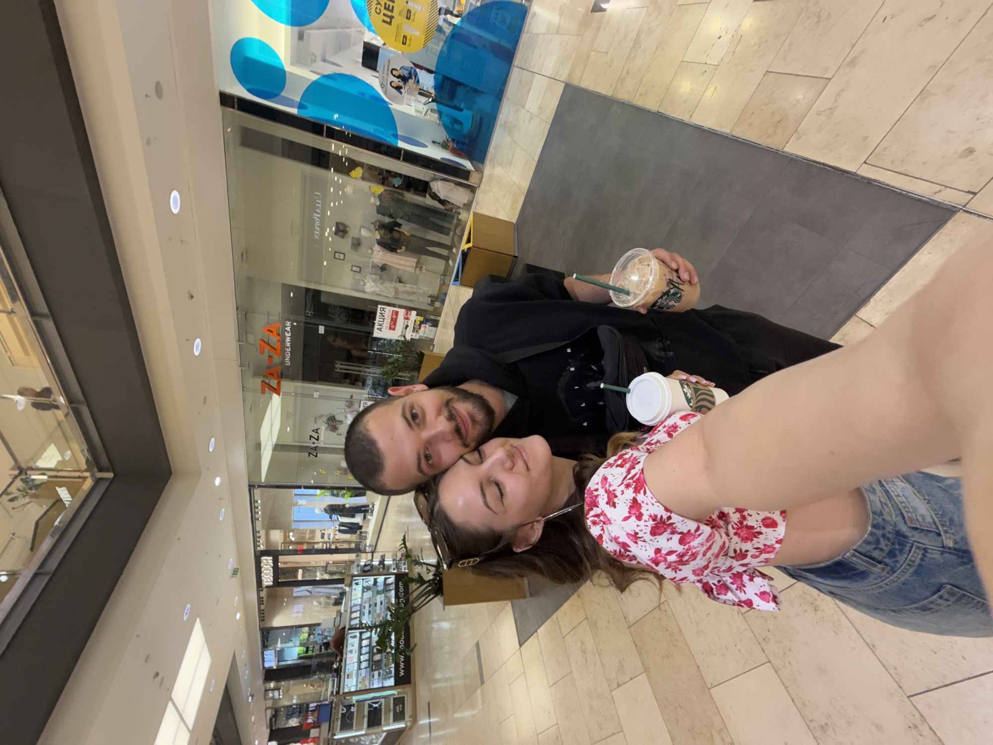 The image shows a man and woman posing for a selfie in what appears to be a shopping mall. The man has a beard and is wearing a black jacket. The woman has shoulder-length hair and is wearing a floral top and blue denim shorts. They are both holding Starbucks drinks. The mall has multiple floors with various shops visible. A clothing store called "ZA-ZA UNDERWEAR" is visible in the background. The setting is likely within a shopping center in Sofia, Bulgaria.