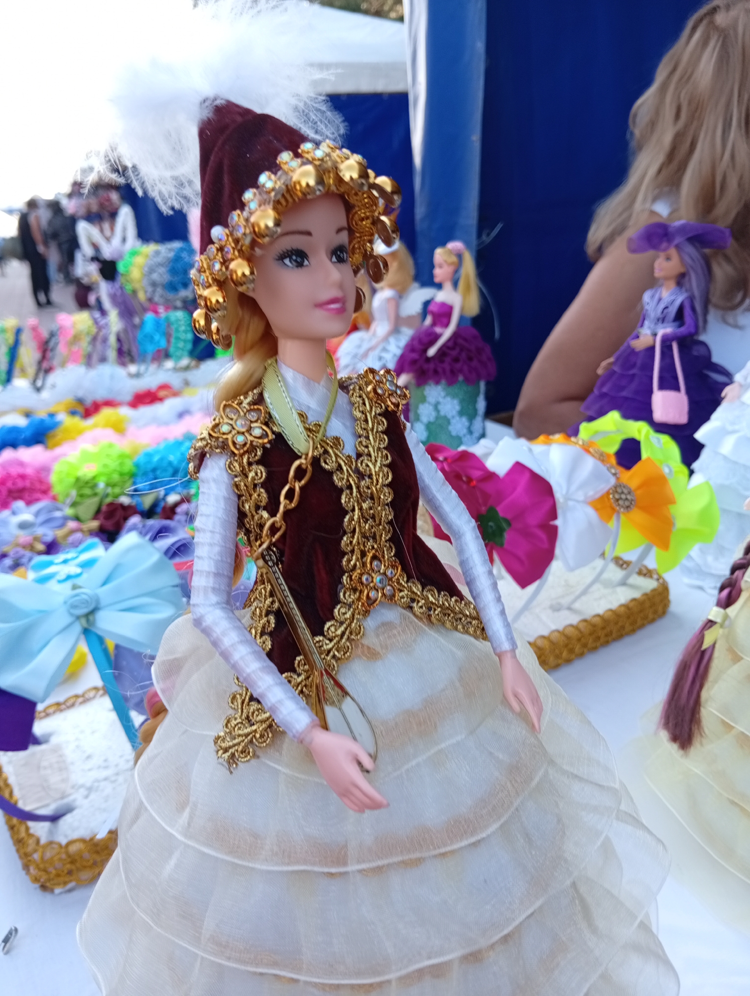 A doll dressed in a traditional maroon and gold vest and a white tiered skirt is displayed outdoors, possibly at a market stall. The doll wears a maroon hat adorned with gold beads and white feathers. Several other dolls and various colorful hair accessories are visible in the background, suggesting a commercial setting. The image appears to have been taken at an outdoor event in Kostanay, Kazakhstan, given the context of the doll's traditional attire.
