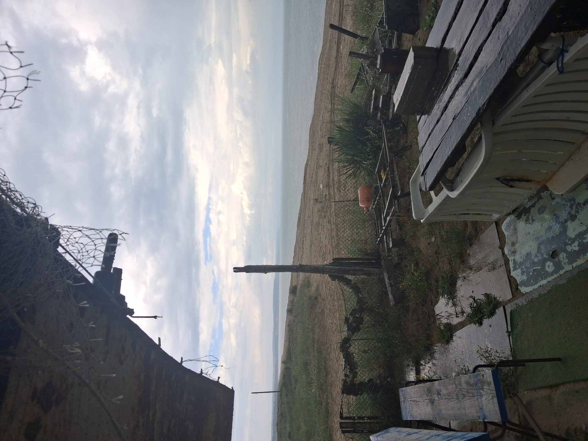 The image presents an outdoor, coastal scene in Shkorpilovtsi, Bulgaria, under an overcast sky. The foreground features damp, uneven ground with patches of sparse green vegetation, exposed soil, and several small puddles of water collected on what appear to be concrete or tile remnants.
In the lower left, a weathered wooden bench with blue-painted metal supports is partially visible. To the right of the bench, a makeshift table is formed by a white, ribbed plastic base (possibly an inverted sun lounger) topped with multiple dark, wet wooden planks. A grey rectangular brick rests on these planks. Adjacent to this table, and extending across the lower midground, various objects are scattered, including an orange, spherical object (possibly a buoy or small gas cylinder), several metallic frames, and unidentifiable dark debris.
A chain-link fence, partially obscured and adorned with dark netting or camouflage material, runs across the midground. Several green, bush-like plants are situated directly behind this fence, particularly towards the right. Two prominent, weathered wooden posts stand upright: one on the left side of the fence line, and a taller, darker post centrally positioned at the fence.
Beyond the fence and vegetation, a sandy bank or dune slopes down towards the Black Sea, which occupies the middle and distant background. The sea surface appears calm and greyish-blue. A faint, low-lying landmass or coastline is visible on the distant horizon, merging with the atmospheric haze.
The sky is predominantly covered by a dense layer of grey, overcast clouds, with some brighter, lighter grey patches visible towards the horizon.
In the upper portion of the frame, an overhanging, dark, and weathered structure, possibly the underside of a roof or boat hull, extends from the left. This structure features visible wires, ropes, and several small, light-colored, tassel-like objects hanging from it. A section of dark netting, resembling a fishing net, hangs from the upper right edge of this overhead structure. A dark, cylindrical object, possibly a pipe or a component, protrudes horizontally from the underside of the structure towards the right.
No individuals or fauna are present in the depicted environment. The wet surfaces indicate recent precipitation.
