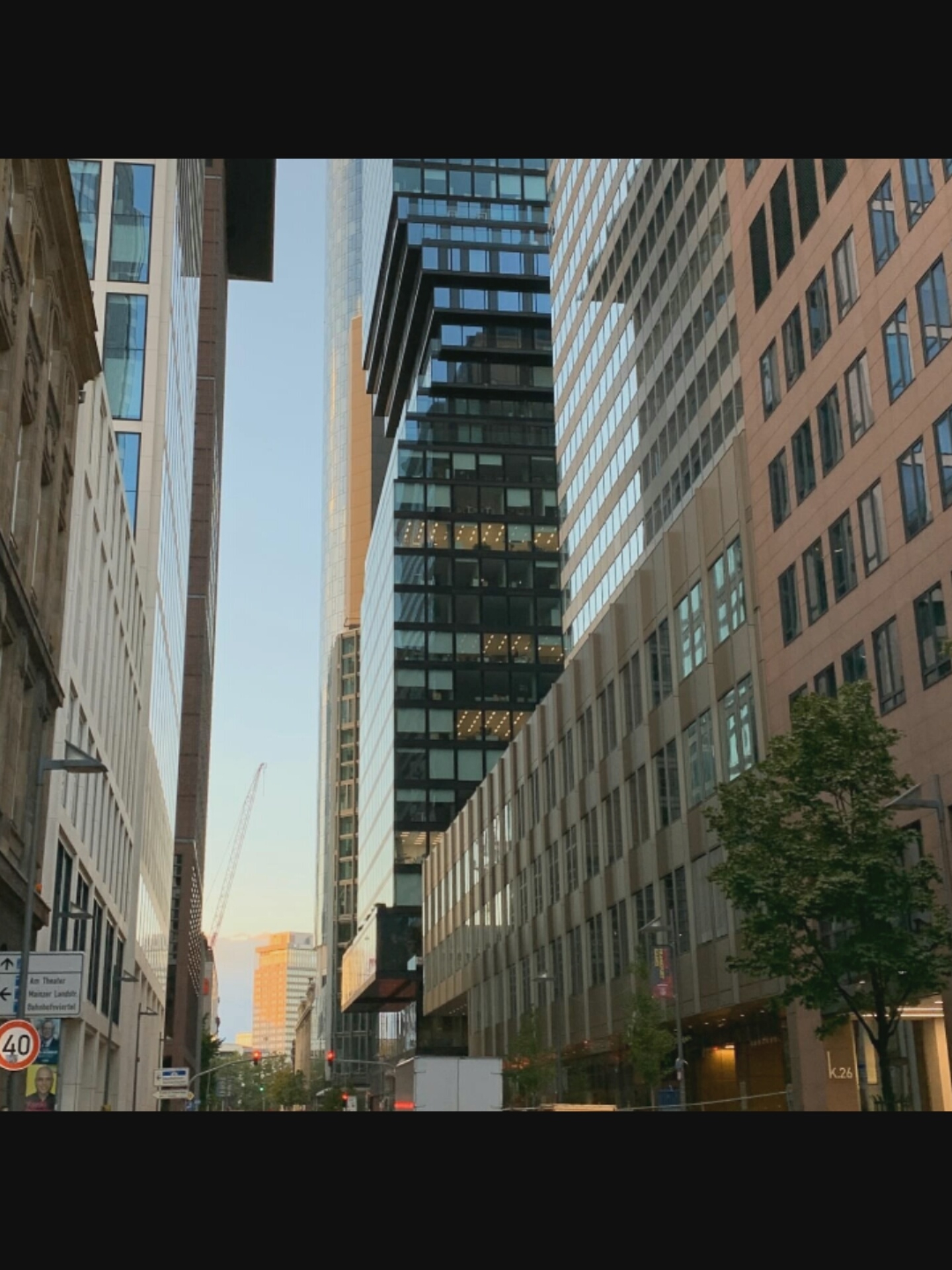 The image shows a street scene in a modern urban environment, likely a business district, with tall buildings on both sides. The architecture suggests a contemporary style with glass facades and geometric designs. A construction crane is visible in the distance, indicating ongoing development. Street signs and traffic signals are present, suggesting a functioning city. One sign has text that appears to be in German, specifically mentioning "Am Theater," "Mainzer Landstr.," and "Bahnhofsviertel," which are indicators of Frankfurt, Germany. There are no people clearly visible in the image. The lighting suggests it is either early morning or late afternoon, with the sun casting a warm glow on some of the buildings. The overall impression is one of a busy, developed urban area.