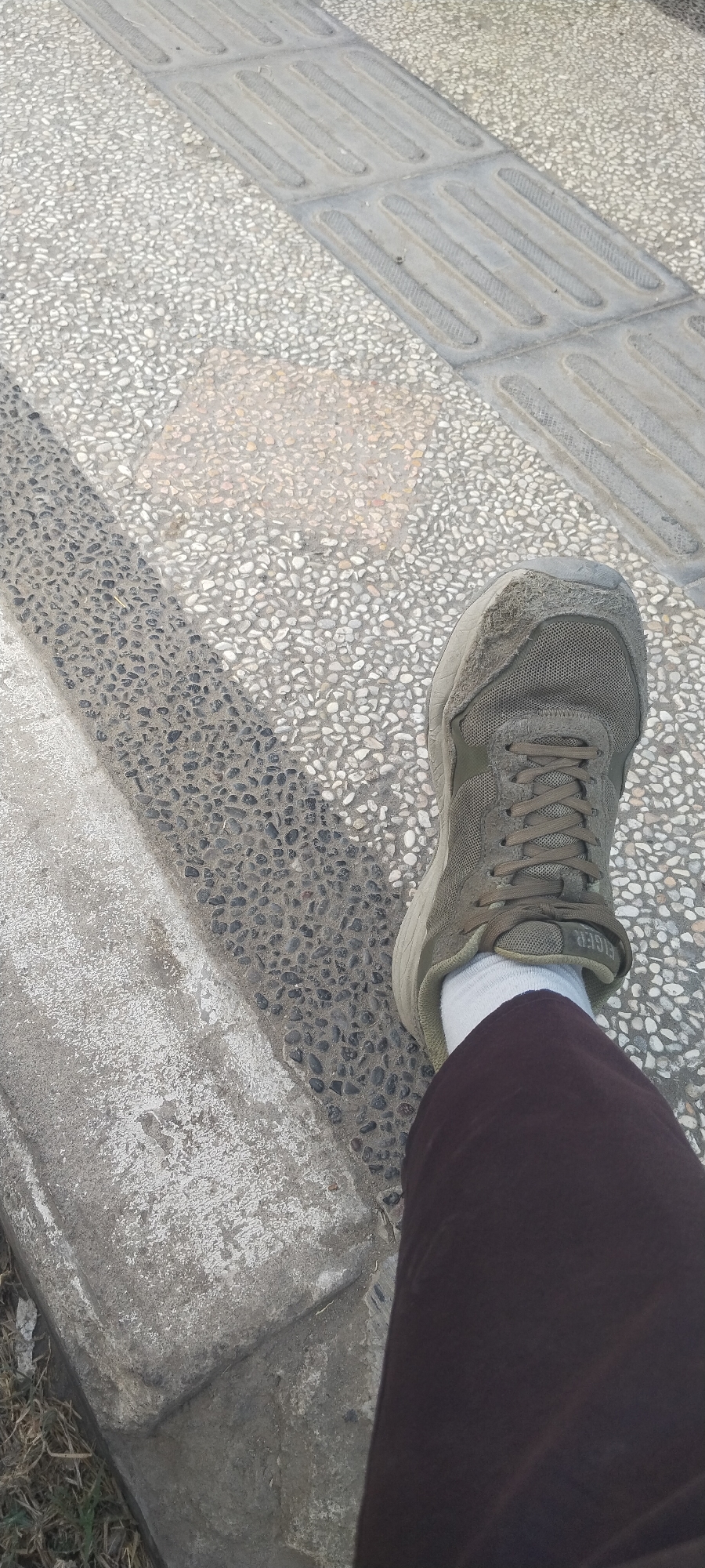 A first-person perspective shot shows a person's right foot wearing a faded olive-green athletic shoe with white socks and dark pants. The shoe is positioned on a sidewalk composed of small, light-colored pebbles. Adjacent to the pebble section is a strip of concrete with raised, parallel lines, suggesting a tactile paving surface for visually impaired individuals. A shadow of the shoe and leg casts diagonally across the pebble-covered ground. To the left of the shoe, a section of a concrete curb is visible, worn and chipped, with some dry grass and debris at its base. The lighting indicates it is daytime, with sunlight creating distinct shadows. The location is specified as Bedah Lawak, Indonesia.