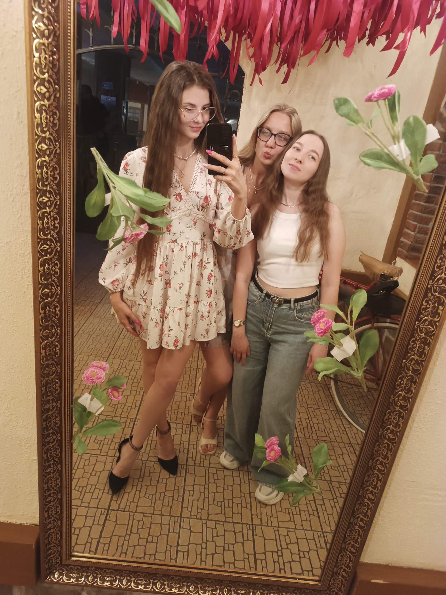 Three young women are reflected in a large, ornately framed mirror. The woman on the left wears a floral dress and black heels, and she holds a cell phone to take the photo. The woman in the center wears a white top and blue jeans. They appear to be indoors, and the mirror reflects a bicycle in the background. The scene is decorated with pink streamers. The city is unknown.