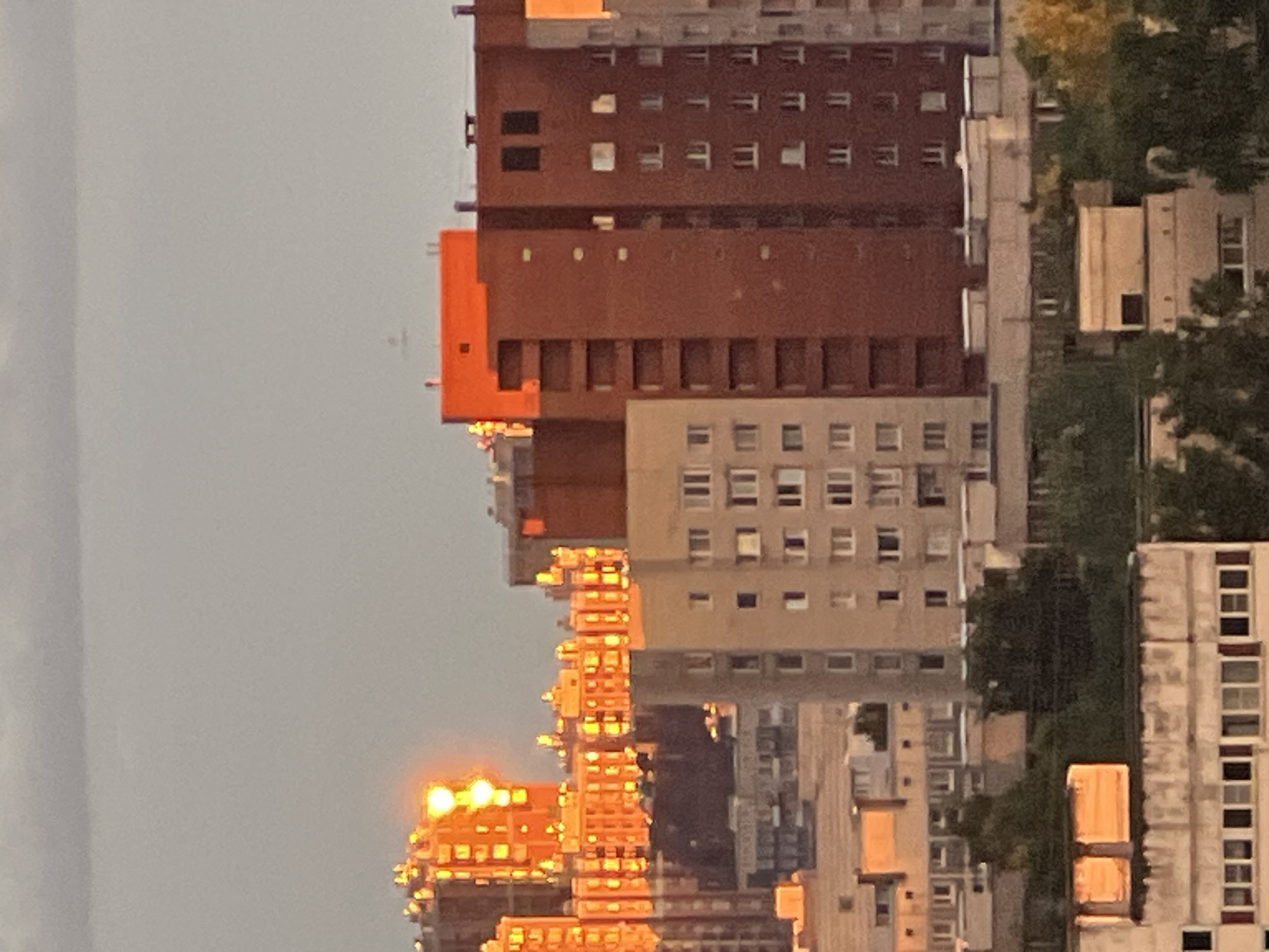 This is a medium-shot, vertical, eye-level, outdoor, photographic image of an urban skyline under a cloudy, gray sky. The sun, partially obscured by clouds, casts a warm, orange glow on several buildings in the distance, creating a dramatic effect. The buildings are a mix of residential high-rises and other structures, predominantly rendered in muted tones of beige, gray, and reddish-brown. Some buildings exhibit a stark, modernist architectural style with repetitive window patterns. The foreground features closer buildings, also residential, with visible windows and architectural details. Green trees are interspersed throughout the scene, particularly in the mid-ground and foreground, suggesting a relatively urban environment with some greenery. The overall impression is one of an urban landscape during either sunrise or sunset, given the lighting conditions. There are no people or animals visible in the image.