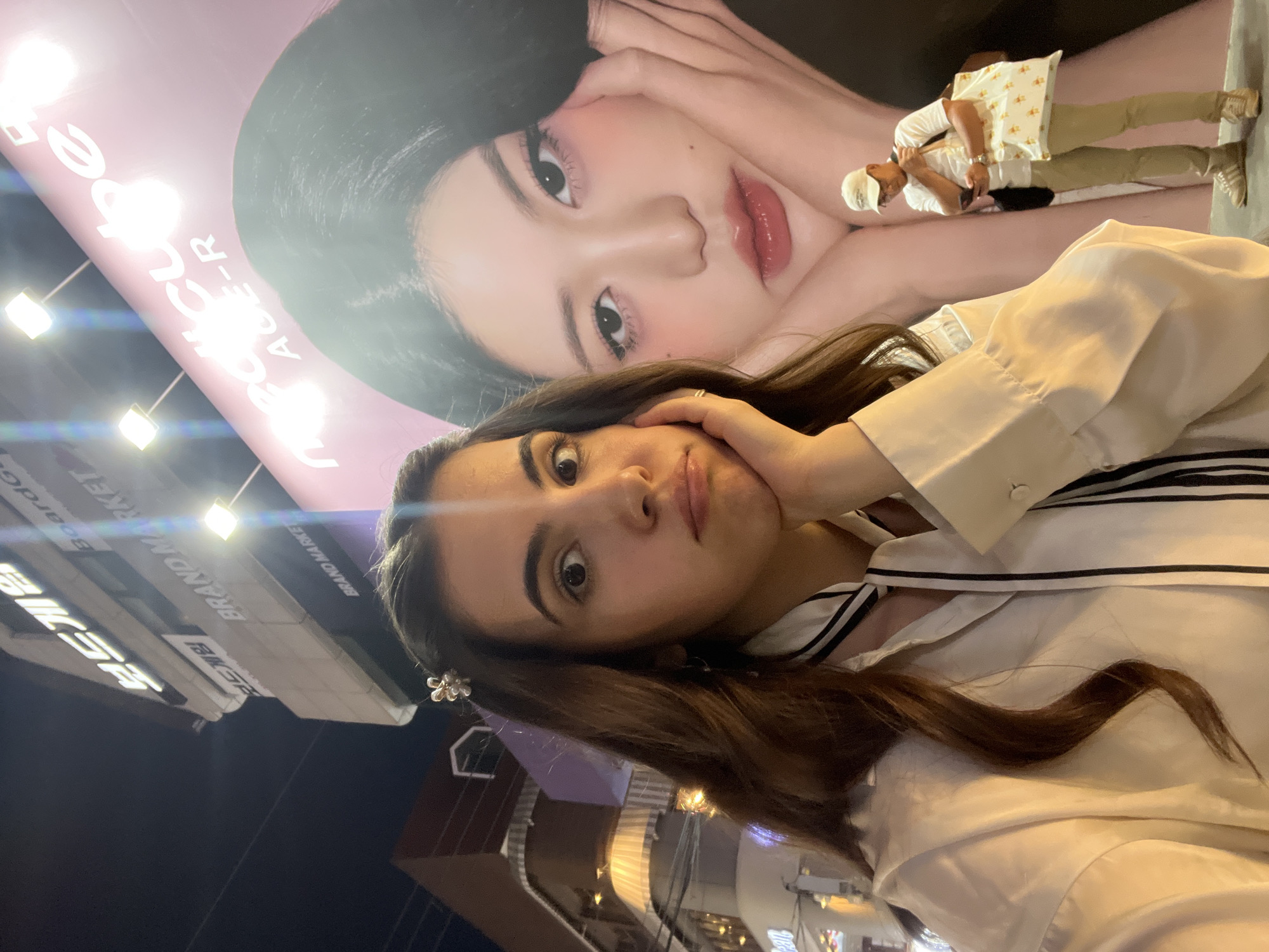 A young woman with long brown hair and a floral hair clip poses for a selfie in front of a large advertisement in Seoul, South Korea. She is wearing a white, collared shirt with black stripes and has her hand touching her face.
Behind her is a large advertisement featuring a fair-skinned woman with dark hair and pink lipstick. A man stands near the advertisement, seemingly looking at his phone.
In the background, a building marked "BRAND MARKET" can be seen, further indicating the location.