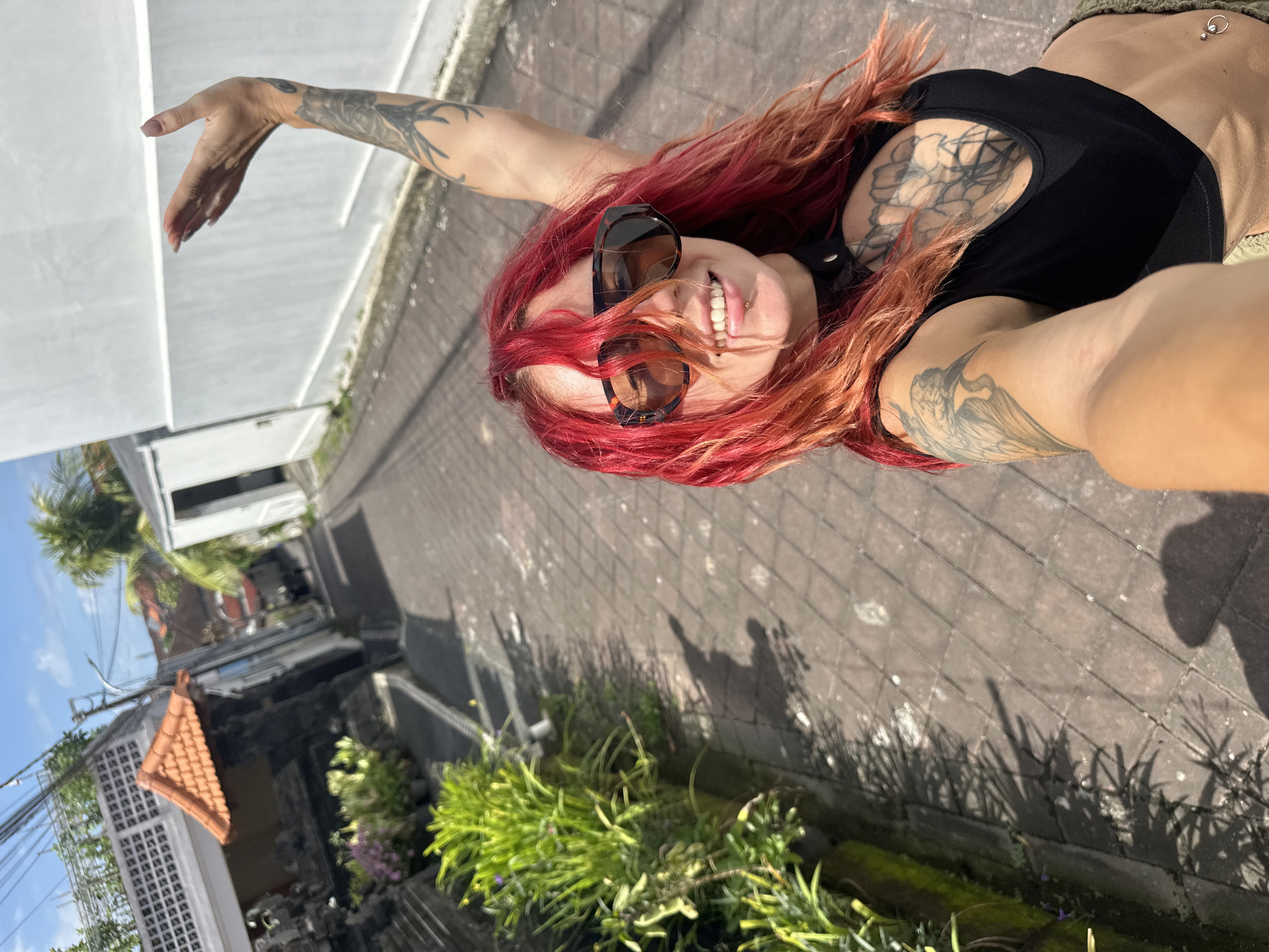 A woman with red hair and multiple tattoos poses for a selfie outdoors. She wears sunglasses and a black top, revealing a navel piercing. Her right arm is outstretched, and she is smiling.
The background shows a narrow street paved with interlocking stones, common in North Kuta, Indonesia. On one side of the street, there's a modern white building. On the opposite side, a traditional Balinese structure with a tiled roof is visible, along with lush green vegetation, suggesting a residential area with tropical landscaping. Overhead power lines are visible against a blue sky. The sun casts distinct shadows.