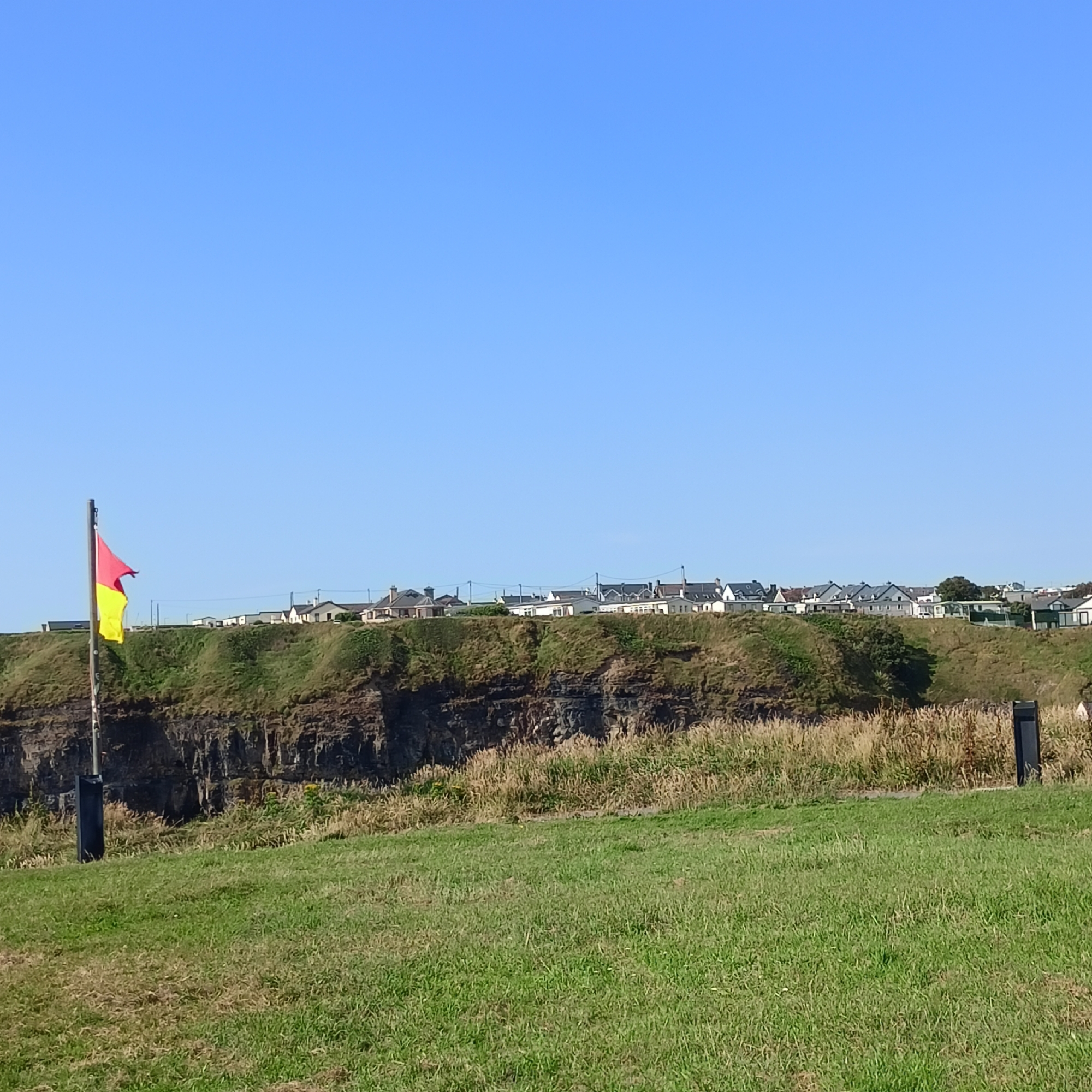 A red and yellow flag on a wooden pole flies on a grassy cliff edge overlooking a coastline. Houses are visible in the distance, situated on top of the cliff. The sky is clear and blue. The scene is a depiction of the coastline near Ballybunnion, Ireland.