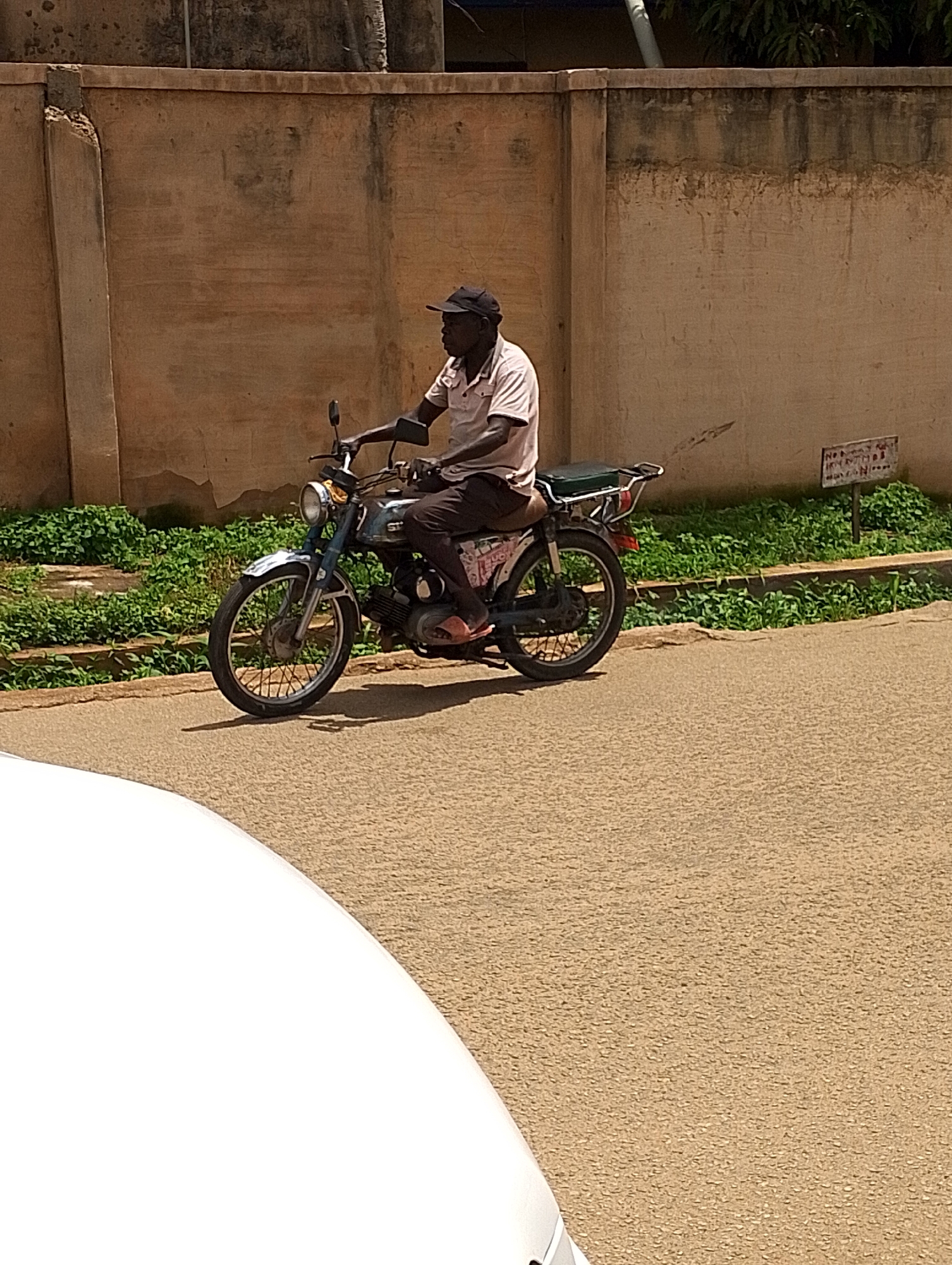 A man is riding a motorcycle on a paved road in Jos, Nigeria. He is wearing a light-colored short-sleeved shirt, dark pants, and a black baseball cap. The man is looking forward, appearing to be focused on the road ahead. The motorcycle is blue with a chrome fender and has a black seat. It appears to be an older model.
The scene is outdoors, with a textured, beige wall in the background. Green foliage is growing along the base of the wall, suggesting a partly vegetated area. A small, weathered sign with illegible text is attached to the wall. The road surface is a textured asphalt. The lighting suggests it is daytime, likely mid-morning or afternoon, with clear weather. A white object, possibly the hood of a car, is partially visible in the foreground, obscuring the lower left portion of the image. The overall impression is a candid snapshot of daily life in Nigeria.