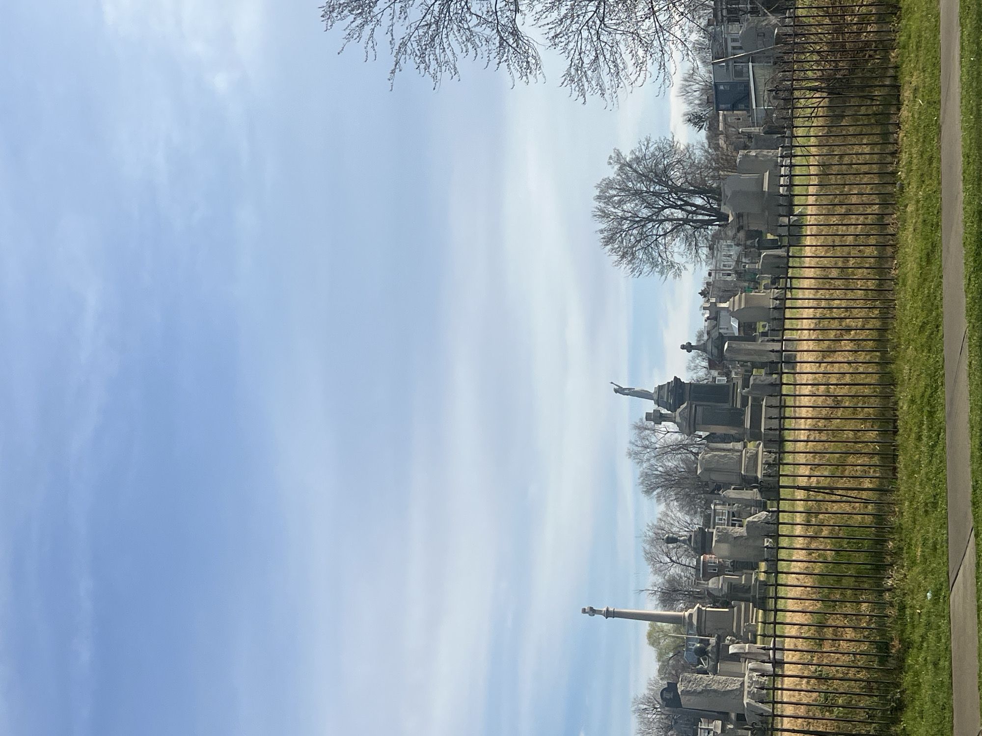 The image shows a daytime view of a cemetery. A wrought iron fence runs along the perimeter, separating the cemetery from a paved walkway and grassy area. Gravestones and monuments of varying sizes and styles are visible within the cemetery grounds. Some bare trees are interspersed among the gravestones. A tall monument and a statue are also visible. The sky is light blue with wispy clouds. The location is likely in the City of Newburgh, United States of America, due to the question's requirement.