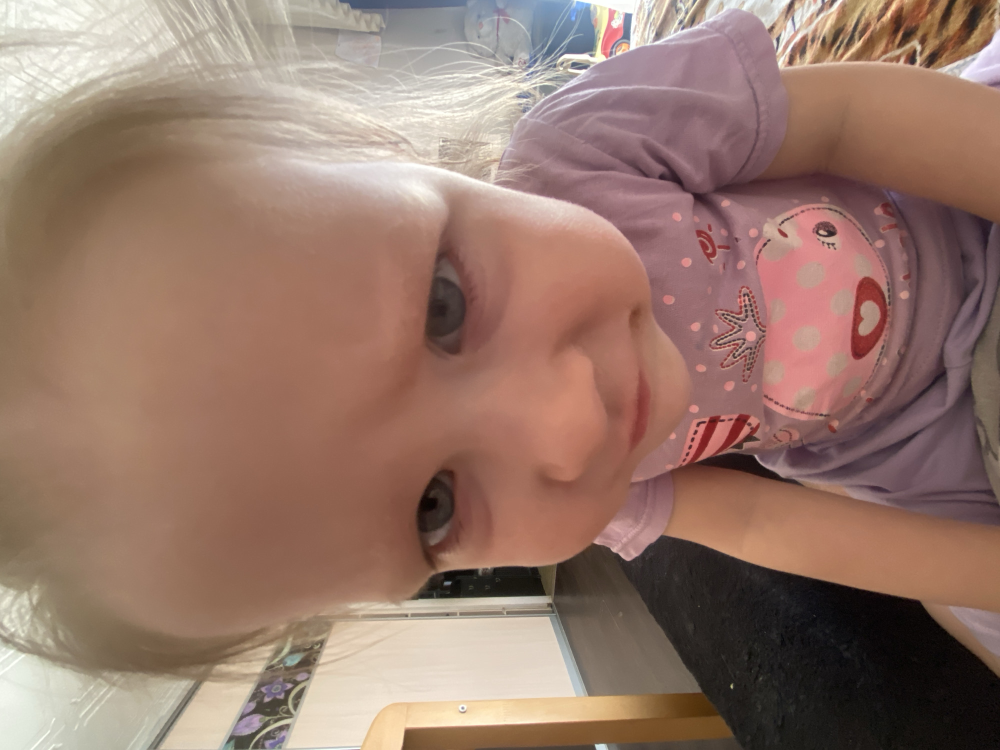 A young child with blonde hair and blue eyes is lying down, wearing a purple t-shirt with a pink cartoon bird design. The background includes a window with blinds, a wooden chair frame, and flooring with a decorative border. The location is Chaikovsky, Russia.