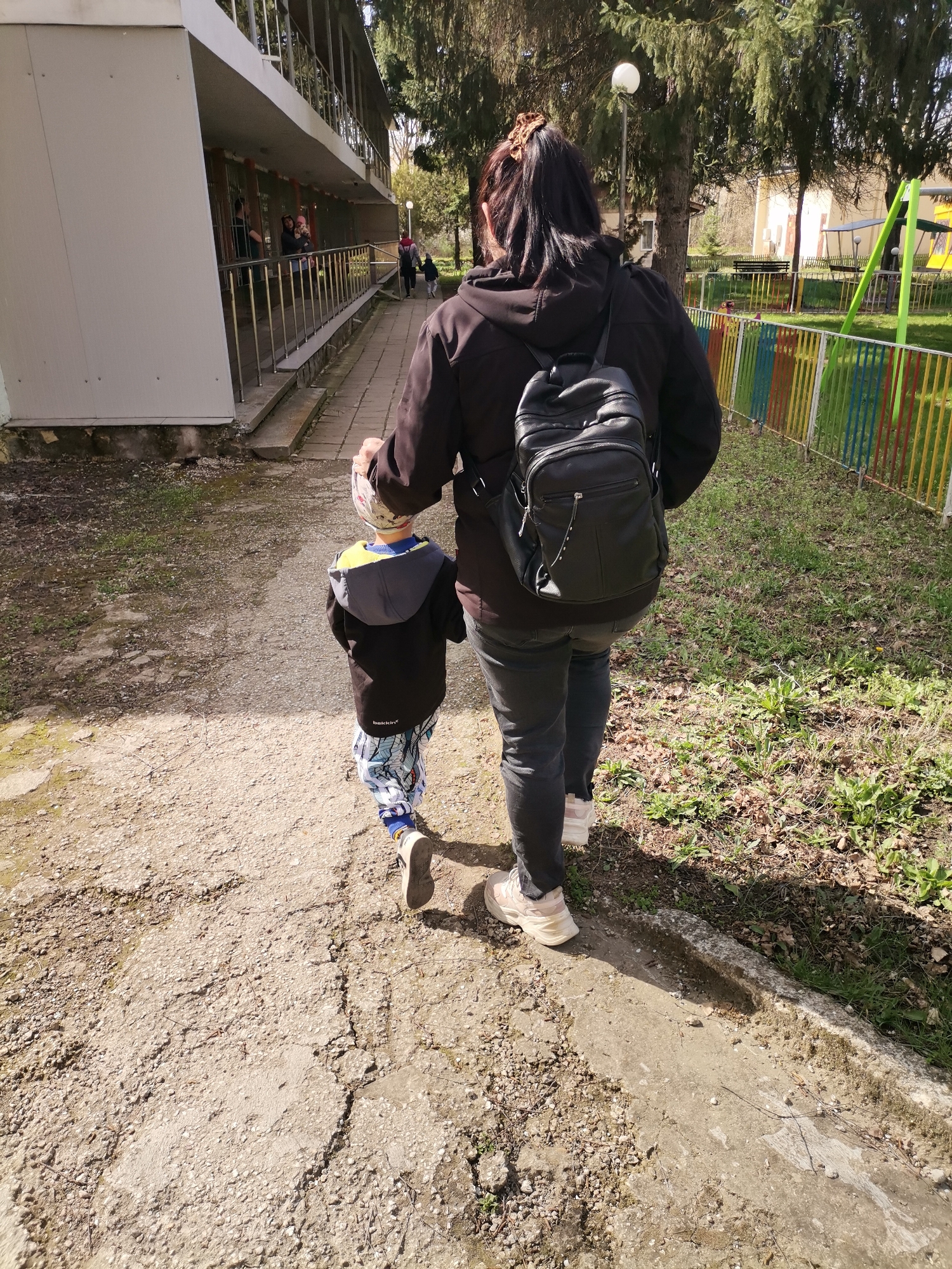 A woman and a young child are walking away from the camera on a cracked concrete path. The woman is wearing a dark jacket, dark jeans, and light-colored sneakers. She has a black backpack on her back. The child is wearing a dark jacket with a yellow hood, patterned pants, and sneakers. They are holding hands. To the right is a grassy area with a colorful fence and playground equipment. To the left is a building with a metal railing. The location is Dobrich, Bulgaria.