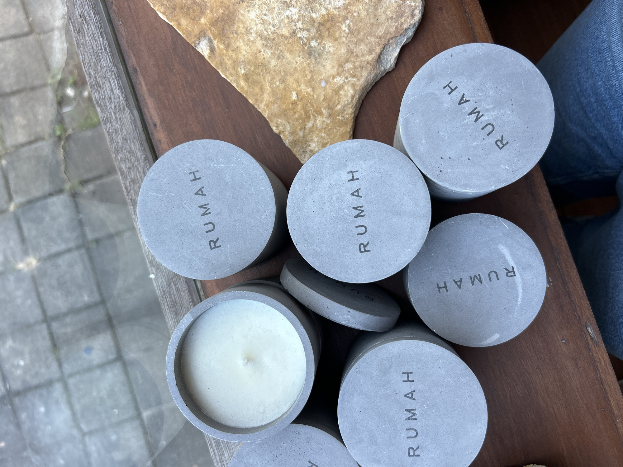 A high-angle medium shot shows a collection of seven gray, cylindrical concrete candles with the word "RUMAH" stamped on their lids, arranged on a dark brown wooden surface. One candle is lit, revealing a white wax interior with a wick. To the right of the candles, a large, flat, tan-colored stone with visible texture sits on the wooden surface. The background is out of focus, but appears to be a tiled outdoor patio seen through a glass pane, suggesting the scene is located in North Kuta, Indonesia, possibly on a windowsill or table overlooking an outdoor area.