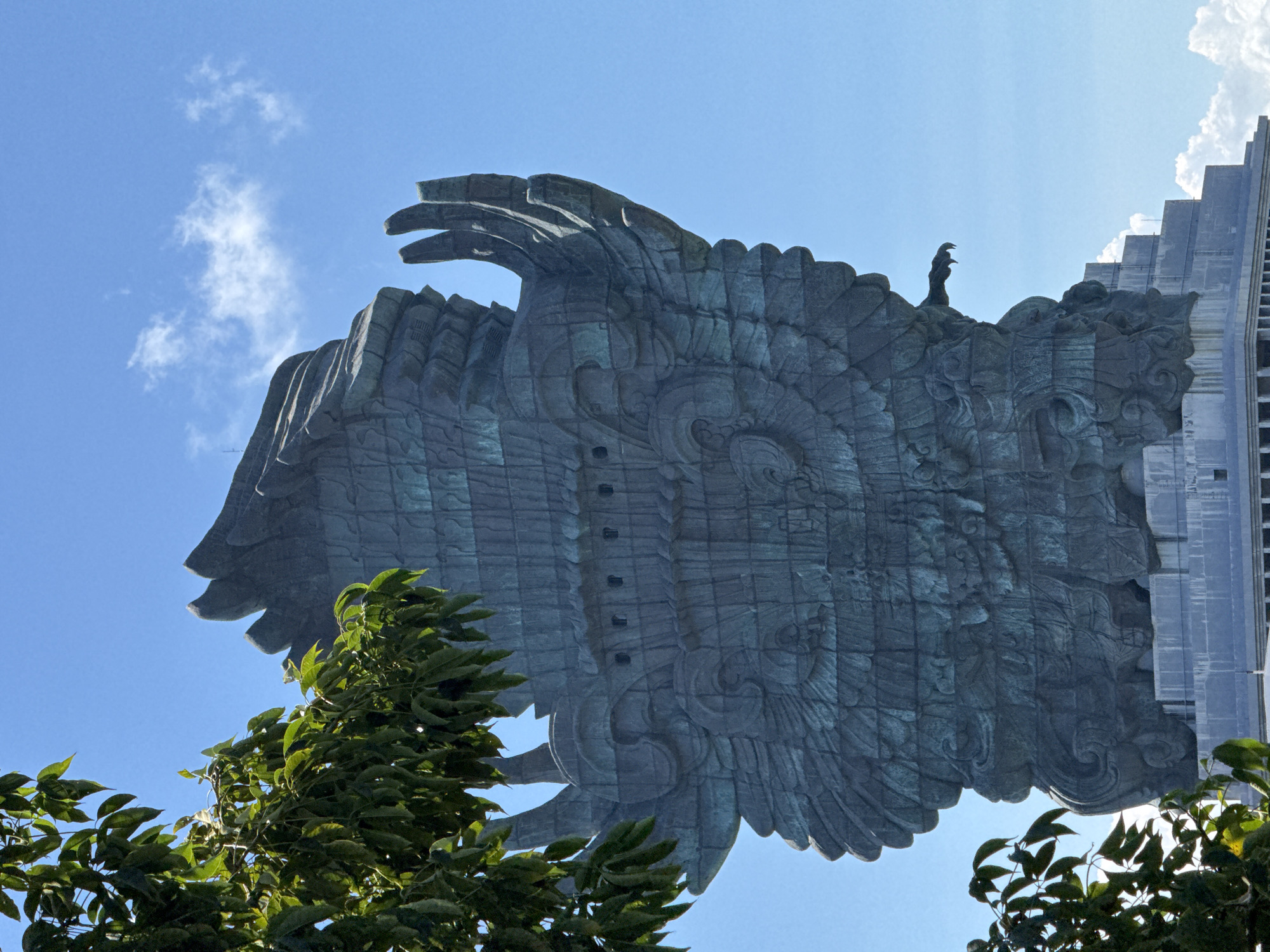 The image depicts a low-angle shot of a large statue of the Garuda Wisnu Kencana, located in Kuta Selatan, Indonesia. The statue's prominent features include intricately designed wings constructed from numerous individual panels. A smaller human figure can be seen standing atop the statue, possibly an observer or maintenance worker. To the right of the statue, a modern architectural structure with a stepped design can be observed. In the foreground, the tops of trees with green leaves partially obscure the lower part of the statue. The sky is a clear blue with sparse clouds.
