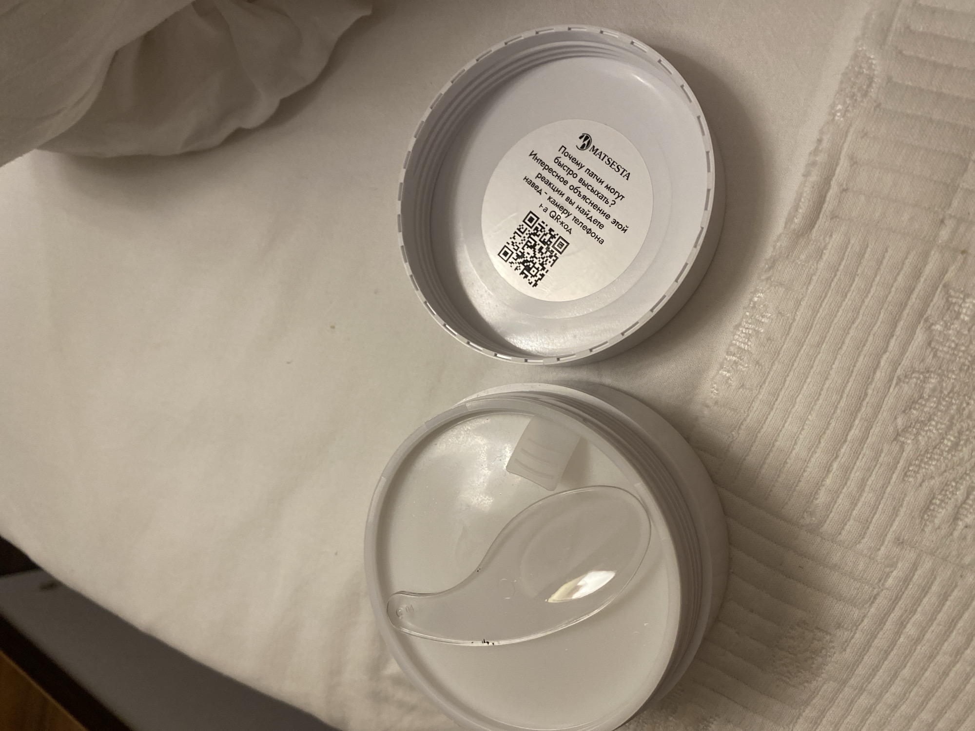 An open white container with a clear plastic spatula rests inside on a white textured surface. The lid of the container is placed next to it, revealing a circular label with a QR code and text in Russian. The text translates to: "Why does this product react? Faster reaction than a phone! Scan the QR code." The brand name "MATSESTA" is also visible on the label. The overall environment suggests a bathroom or vanity area. There are no people present. The location is not specified in the image or provided in the prompt.