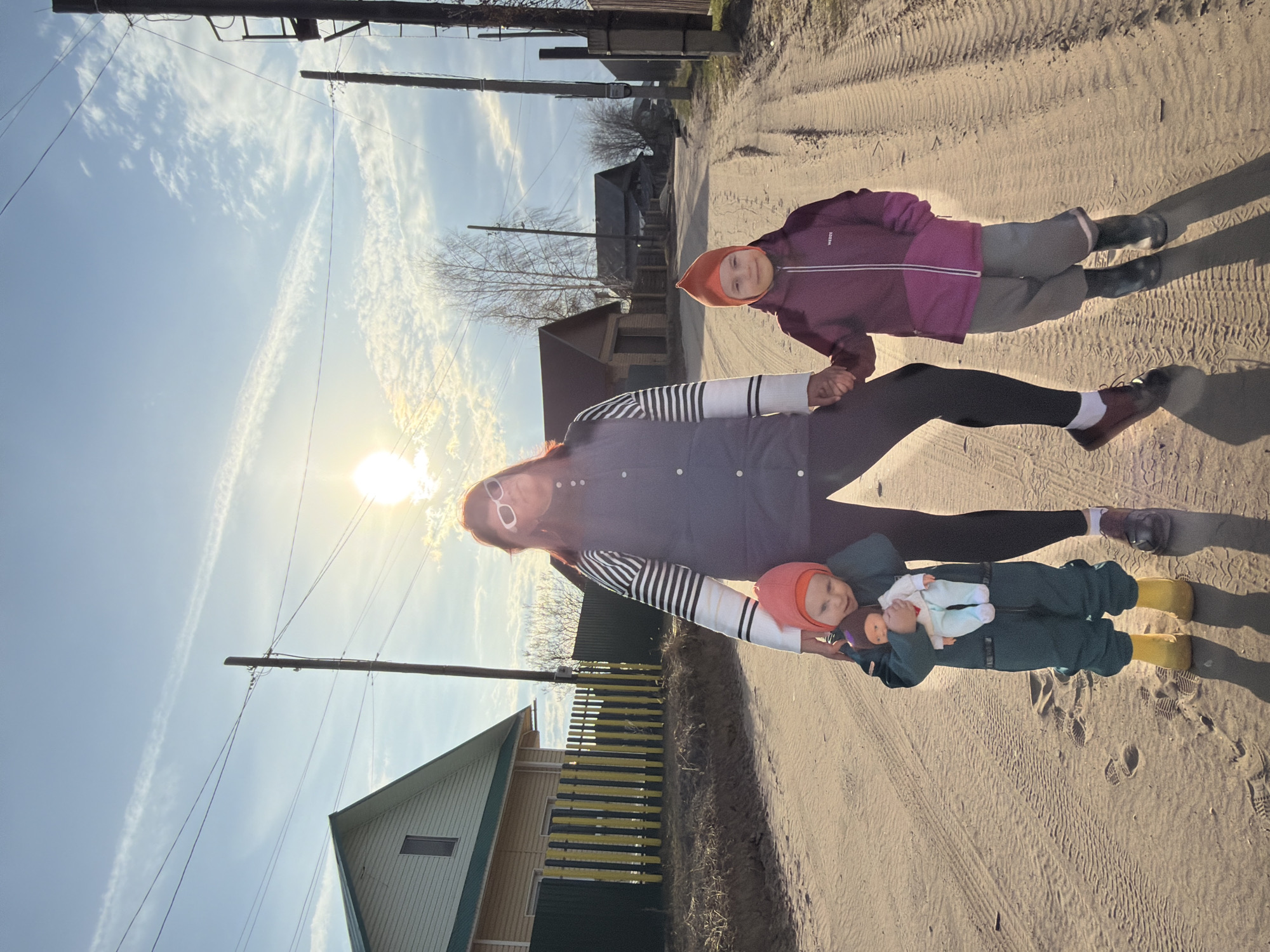 This image depicts an adult and two children walking on an unpaved dirt road under a bright sky in what appears to be a rural or semi-rural setting, consistent with Chaikovsky, Russia. The road shows clear tire tracks and footprints.
An adult, with reddish hair and wearing white sunglasses, a striped long-sleeved shirt, and a dark vest, is holding hands with an older child and carrying a younger one. The older child, dressed in a purple jacket (with "WEDZE" visible on the chest), gray pants, black boots, and an orange hat, looks towards the camera with a calm expression. The younger child, in a green or teal overall suit, yellow boots, and an orange hat, is being carried and holds a doll with a dark head and white clothes.
The bright sun is positioned low in the sky, creating a strong lens flare and casting long shadows of the figures behind them, indicating either early morning or late afternoon. The sky is clear blue with scattered white clouds and distinct contrails. In the background, wooden houses, some with fences, and bare trees are visible, alongside utility poles and power lines, reinforcing the rural character of the scene. The overall atmosphere appears pleasant and relaxed.