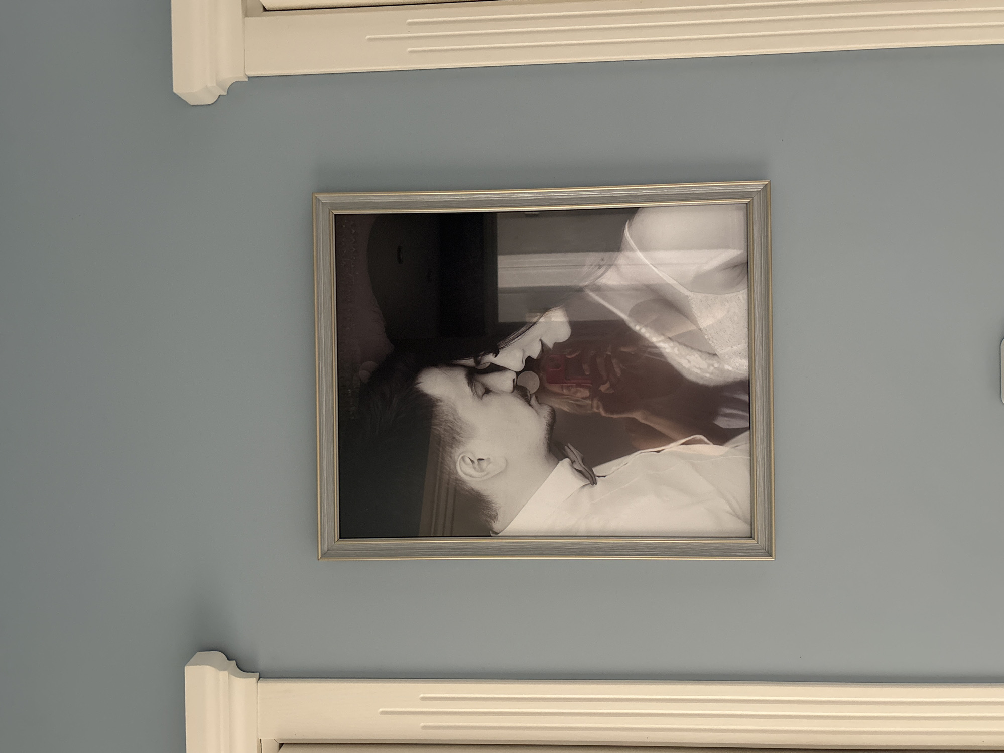 A silver-framed black-and-white photograph hangs on a blue wall. The photograph depicts a kissing couple. Framing the top and bottom of the photograph are sections of white molding.