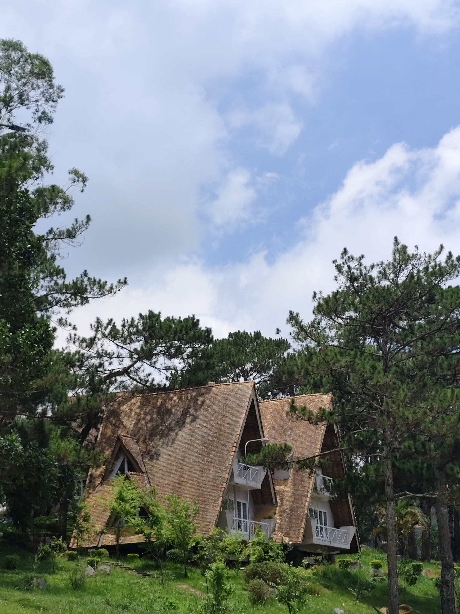 The image shows a unique architectural structure nestled among pine trees, with a backdrop of a partially cloudy sky. The building has a distinctive A-frame design with tiled roofs and white balconies. The structure is elevated from the ground, with green foliage and trees surrounding it. The location is likely in Da Lat, Vietnam, which is known for its pine forests and distinctive architecture. There are no people visible in the frame. The environment suggests a serene, natural setting with a building that blends into its surroundings.