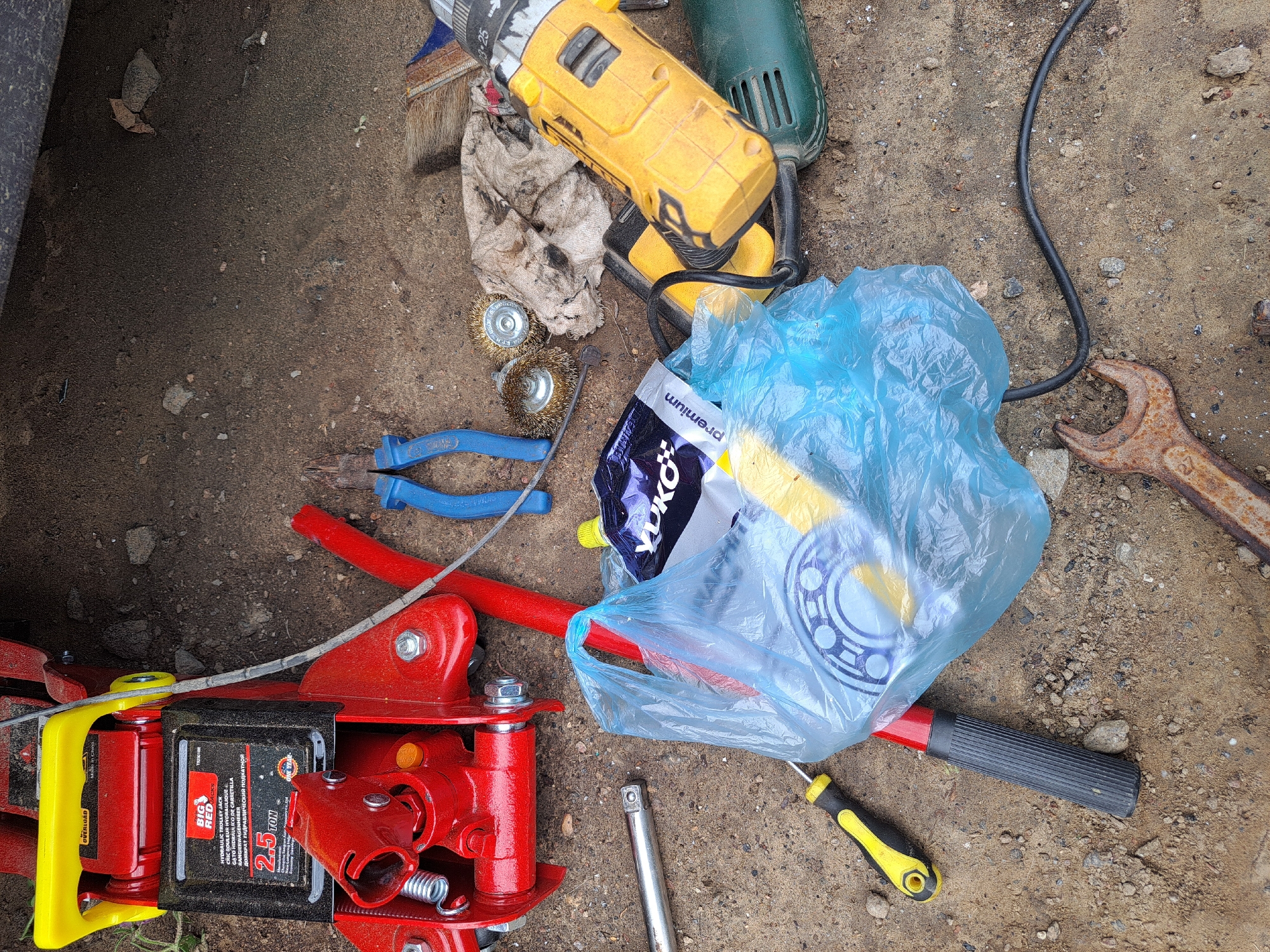 The image depicts a collection of tools and equipment scattered on a dirt ground, likely in an outdoor setting. A red hydraulic trolley jack with yellow accents is prominent in the upper left. Adjacent to it are a pair of blue-handled pliers and a silver socket wrench. In the center, a blue plastic bag contains various items, including what appears to be a bearing and some yellow packaging with "YUKO premium" visible. To the right of the bag, a yellow and black screwdriver lies on the ground. Further to the right, a yellow power drill and a green grinder are visible. Several wire brushes and a dirty rag are also present. In the bottom right corner, a rusty wrench is partially visible. The ground is composed of dirt, small stones, and some dry leaves. The overall scene suggests a work area where repairs or maintenance may have been performed. The location is specified as Vypasne, Ukraine.